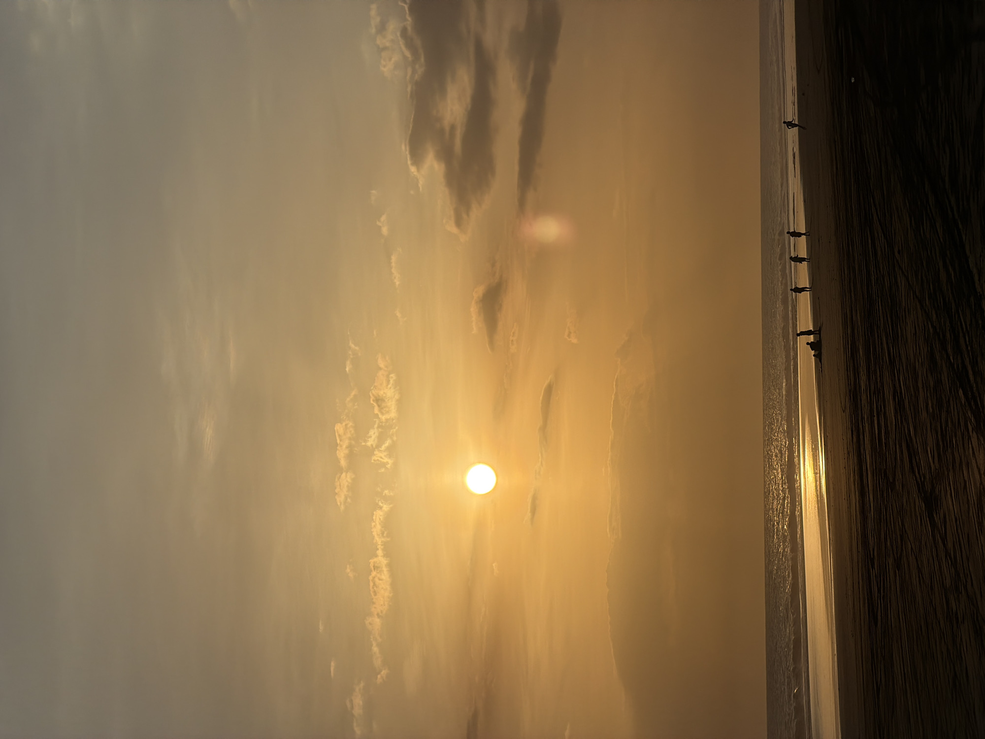 The image depicts a beach at sunset, likely in Selemadeg Timur, Indonesia. The sun is positioned near the center of the frame, partially obscured by clouds, casting a warm golden light across the sky and sea. Silhouetted figures are walking along the shoreline. The sand appears dark, possibly wet, with wave patterns visible.