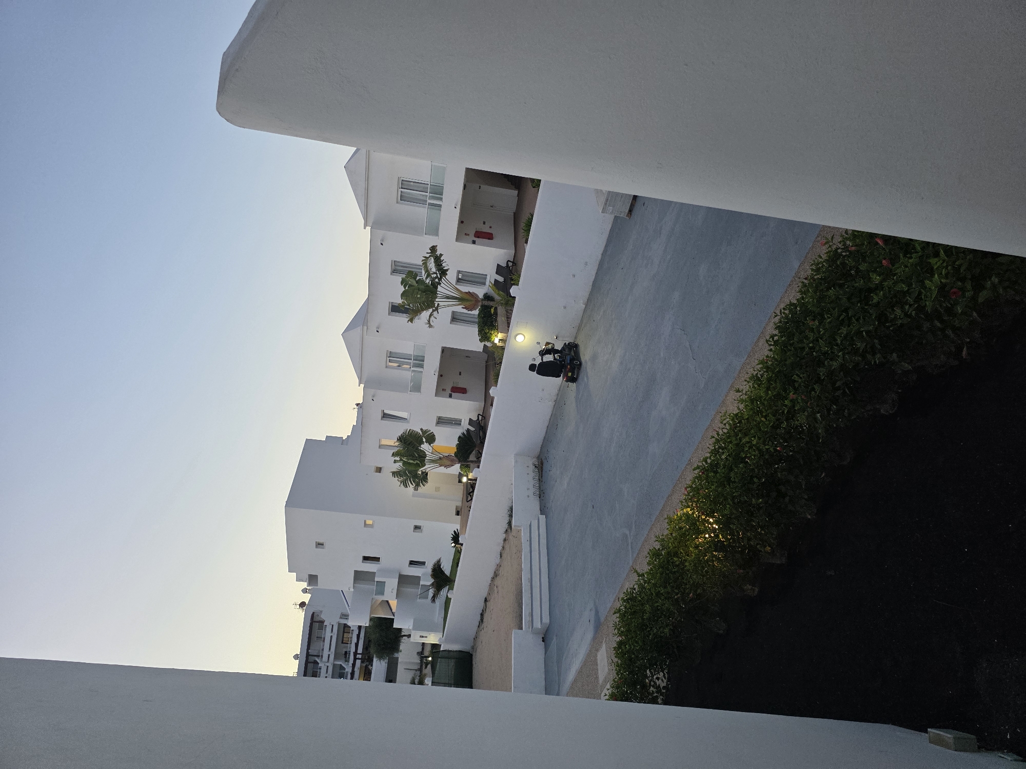 In Costa Teguise, Spain, at dusk, an individual is operating a mobility scooter in a paved courtyard area. White stucco buildings with sharp architectural lines and flat roofs dominate the background, featuring multiple levels, balconies with glass railings, and doorways. Tropical palm trees are interspersed among the buildings. A low white wall encloses the courtyard, with a small set of stairs leading up to a patch of sand. A manicured green hedge runs along the bottom edge of the courtyard. The sky is a pale, clear blue, transitioning into a soft yellow hue near the horizon, indicating the setting sun. The lighting suggests it is late evening.