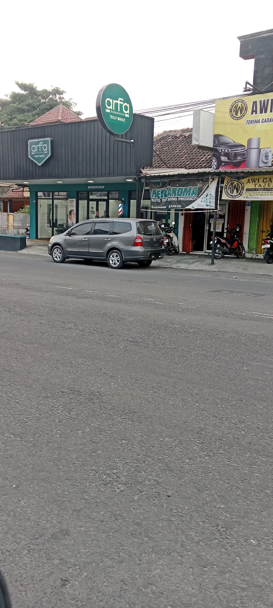 The image shows a street scene in Maguwoharjo, Indonesia, featuring several businesses. On the left, a barbershop named "Arfa Barbershop" is identifiable by its prominent green circular sign and a smaller rectangular sign. Next to it, a grey minivan is parked. To the right of the barbershop, a banner for "FERAROMA AUTO DETAILING PRODUCT" is visible, along with the text "PT AWIGAMA JOGJA" on a yellow sign. This yellow sign also displays images of a car, a refrigerator, and a washing machine. The businesses are situated along a paved road, with motorcycles parked in front of some establishments. The sky is overcast.