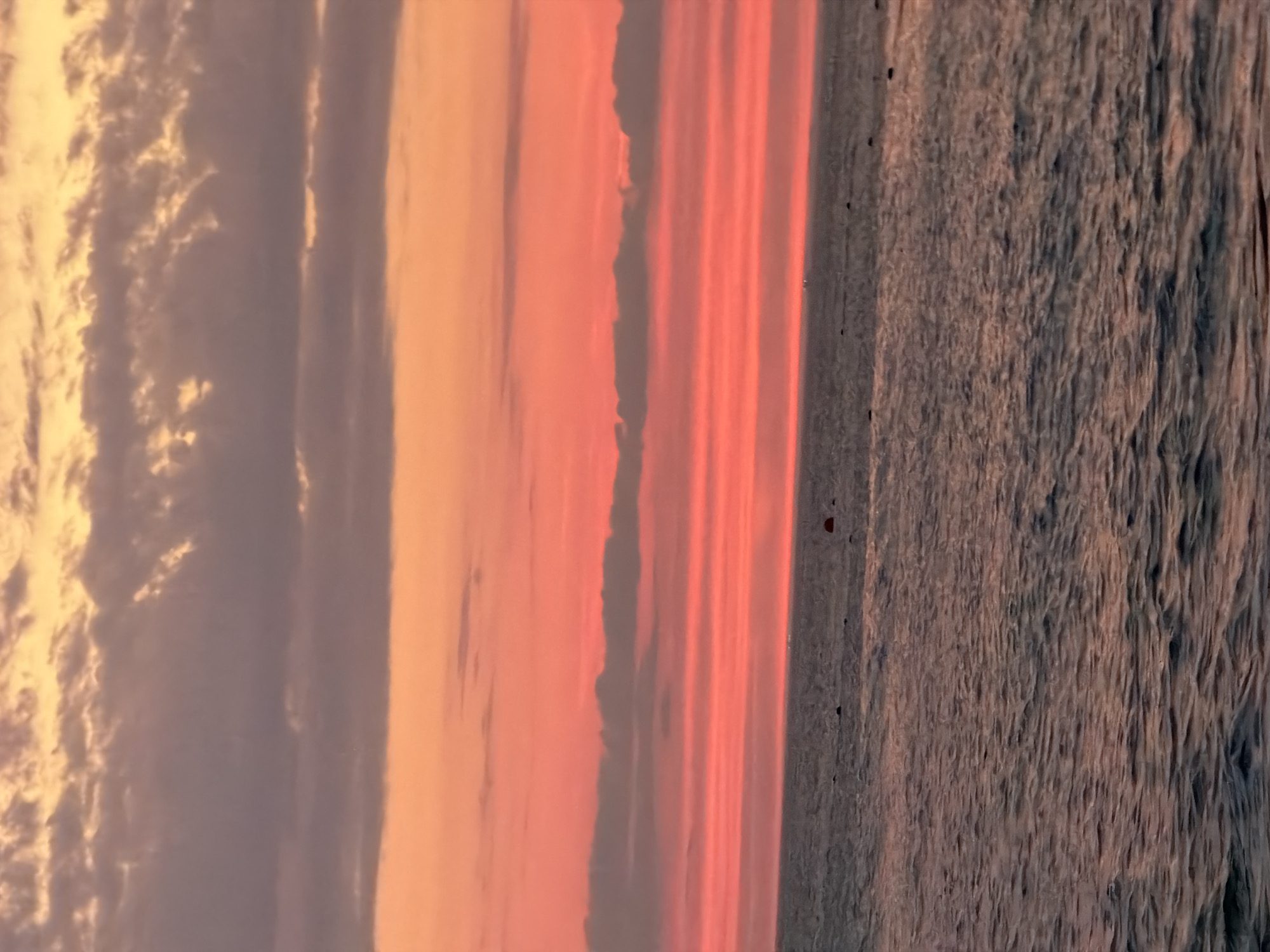The image depicts a serene seascape at sunset, likely near Sochi, Russia. The sky is a vibrant canvas of colors, transitioning from warm oranges and pinks near the horizon to cooler purples and grays higher up. Wispy clouds are scattered across the sky, catching the fading light. The ocean below is calm, with gentle waves reflecting the colors of the sunset. In the distance, a few dark shapes, possibly buoys or small boats, are visible on the water. The overall impression is one of tranquility and natural beauty.