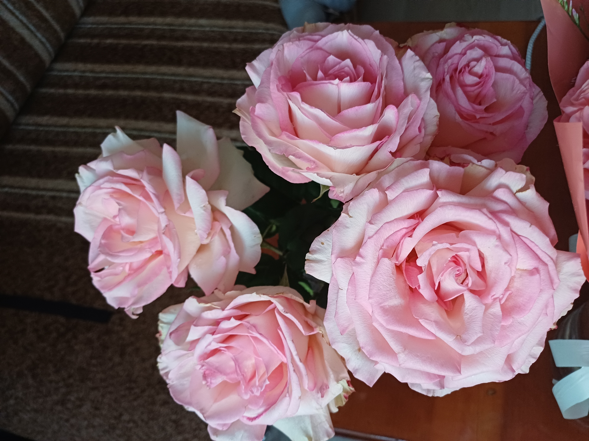 A bouquet of light pink roses is arranged on a wooden surface, with a textured brown and striped background. The roses are in various stages of bloom, with delicate petals unfurling. The lighting is soft, highlighting the subtle variations in color and texture of the flowers. There are no people or animals present in the image. The scene appears to be indoors, possibly on a table or other piece of furniture. The background suggests a domestic setting, perhaps a living room or dining room. There is a hint of a striped fabric, possibly a sofa or curtains, and a dark, possibly wooden, surface. The overall impression is one of natural beauty and simple elegance.