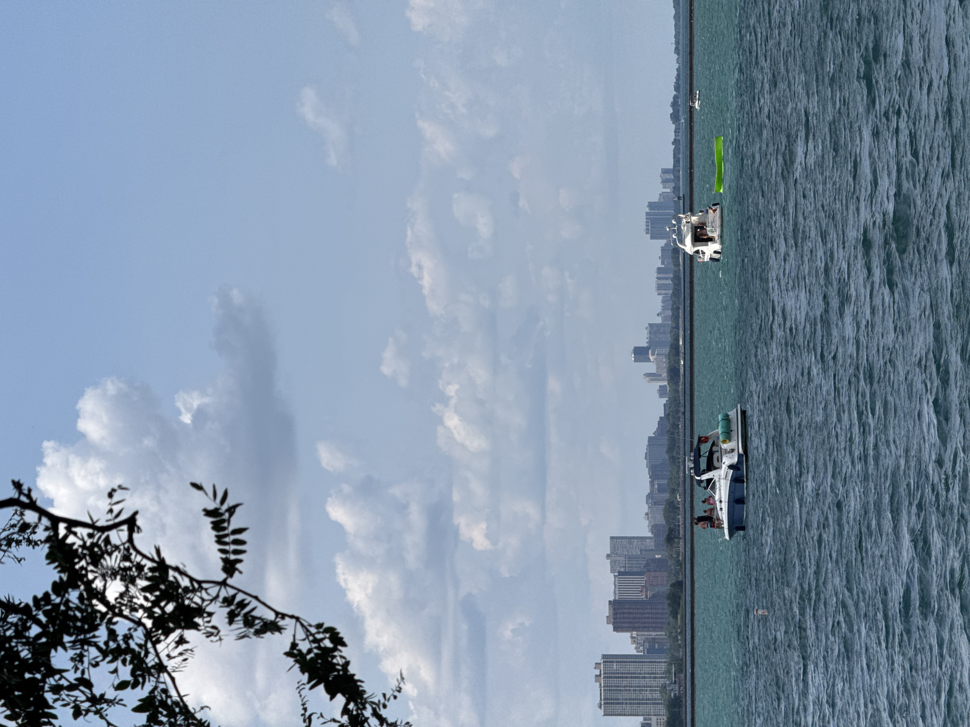 This image captures a view of Lake Michigan with the Chicago skyline in the background. In the foreground, dark tree branches frame the left side of the scene. The water is a deep blue-green, with visible ripples and waves.
On the water, two boats are present. The boat on the left is a white pontoon boat with a blue trim, carrying at least four people who appear to be enjoying a day on the lake. It is positioned in the middle ground of the image. To the right of this boat, a bright green inflatable float is visible on the water.
Further to the right and slightly closer to the horizon, a larger white yacht is seen with several people on board. This yacht is more luxurious in appearance. In the far distance, several other smaller boats are visible.
The Chicago skyline is a prominent feature in the background, stretching horizontally across the image. It consists of numerous tall buildings of varying architectural styles, indicating a dense urban environment. The sky is a bright blue, dotted with scattered white cumulus clouds. The overall atmosphere suggests a pleasant day for boating and leisure on the lake.