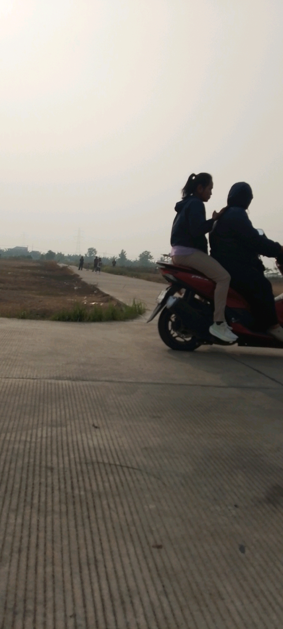 Two individuals are silhouetted on a red motorcycle traveling on a paved road. The person in front, appearing to be a male wearing a dark hoodie, is operating the motorcycle. The person behind, seemingly a female with a ponytail, is wearing a dark hoodie and light-colored pants, with one leg resting on the motorcycle. The road surface features parallel grooved lines. In the background, a hazy, overcast sky hangs over a rural landscape with dry vegetation, scattered trees, and distant buildings, suggesting an outdoor environment in a semi-urban or fringe area of Satriamekar, Indonesia. Several other silhouetted figures are visible in the distance, walking on a path adjacent to the road.