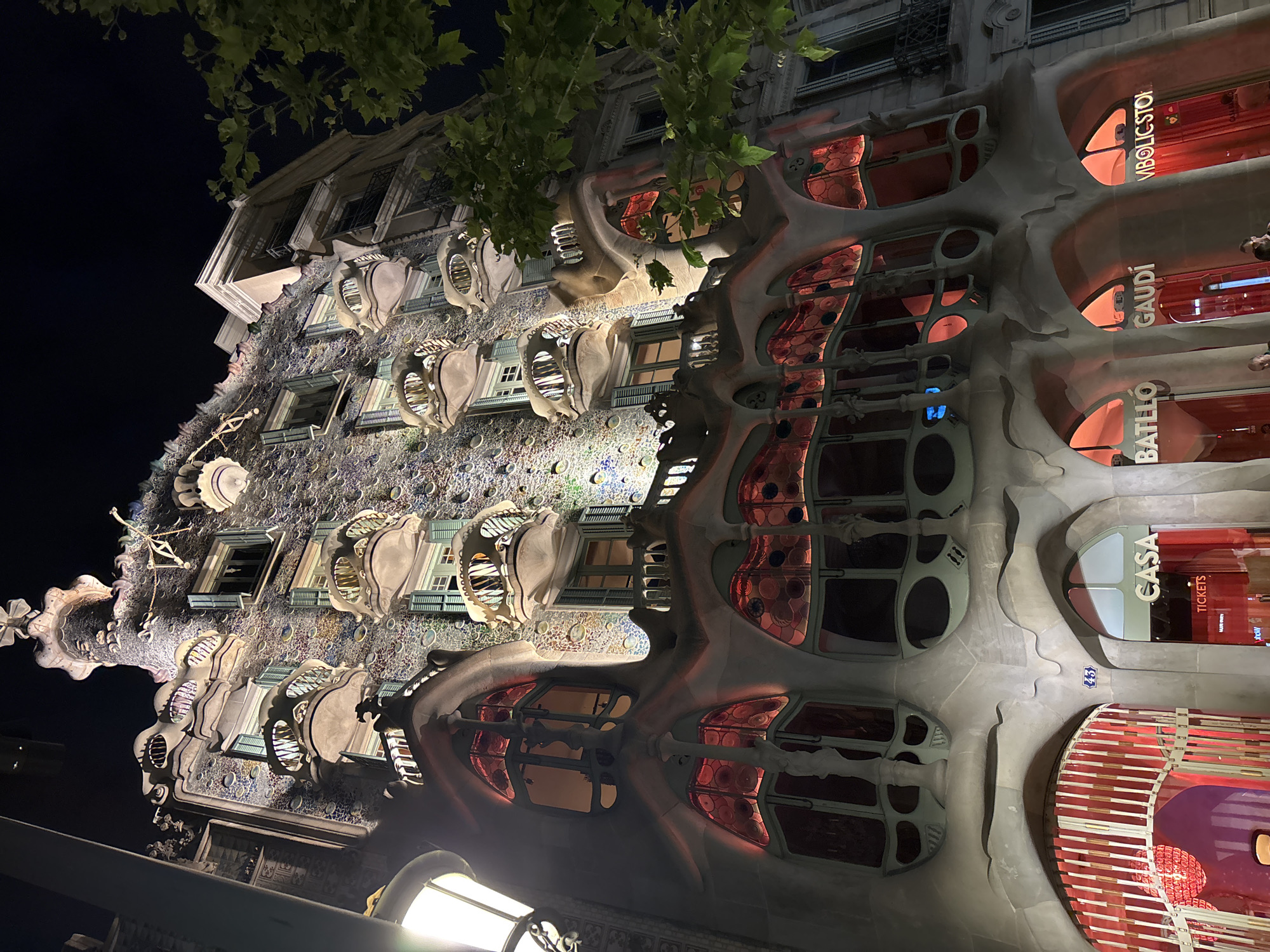 This is a low-angle, night-time, outdoor photograph of Casa Batlló, located on Passeig de Gràcia in Barcelona, Spain. The building's facade is illuminated, highlighting its distinctive organic, undulating architecture and colorful mosaic tiles. Balconies with bone-like structures and iridescent glass windows are visible on multiple levels. The lower portion of the building features arched entrances with red lighting and signage that reads "CASA," "BATLLÓ," "GAUDÍ," and "IMBOLICSTOL." A street lamp is partially visible on the left side of the frame, and a tree with green leaves is on the right. The sky is dark, indicating it is nighttime. No specific actions or interactions of people are clearly discernible.