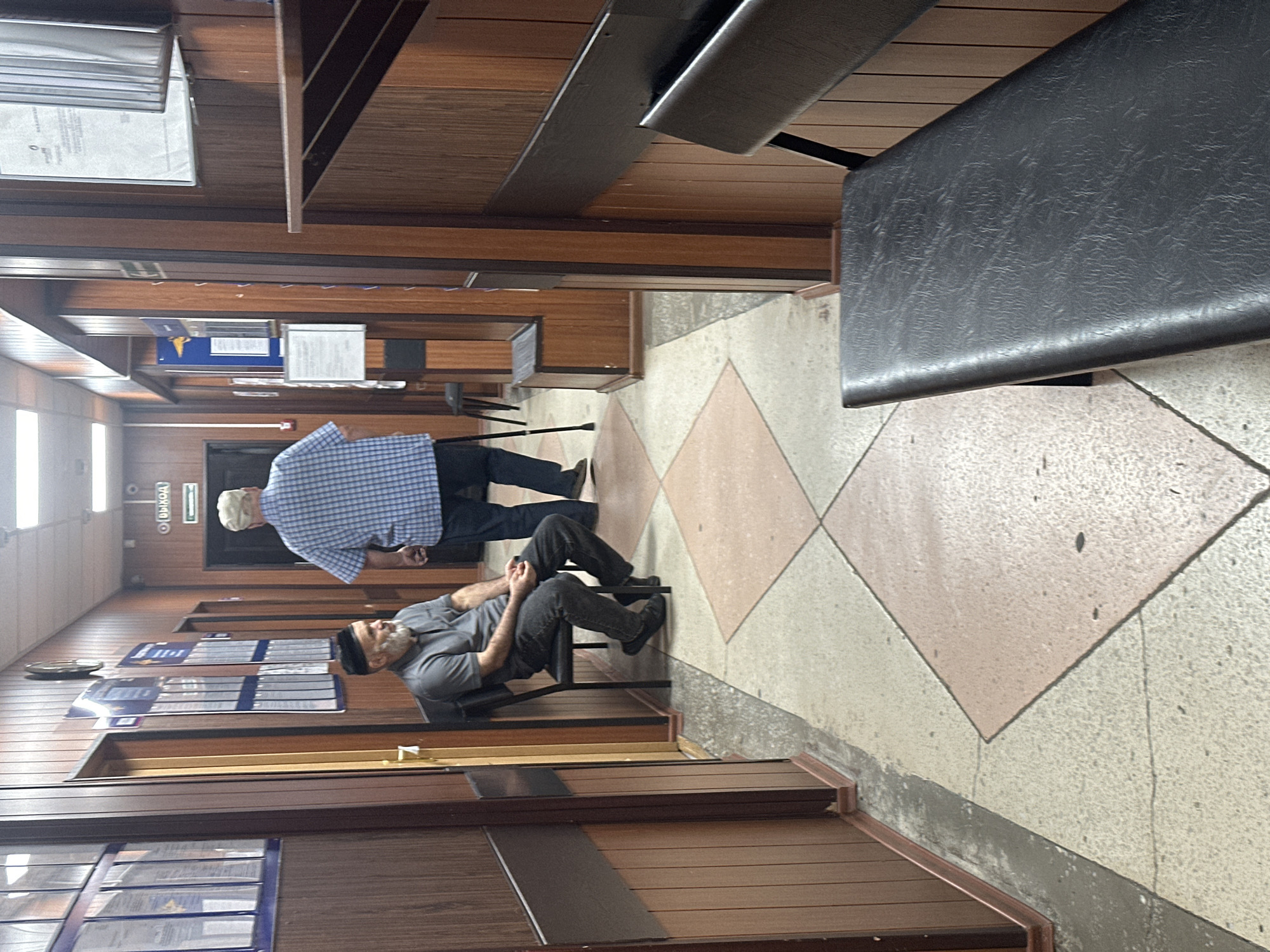 The image depicts a hallway inside a building in Novoaltaysk, Russia. The hallway is paneled with dark wood and has a patterned floor composed of light-colored and pinkish-tan diamond shapes.
Two men are present. In the foreground, to the left, a man with a gray beard, wearing a dark skullcap and a gray polo shirt with dark pants, is seated on a black chair. His hands are clasped in his lap. Further down the hallway, a taller man wearing a blue and white checkered shirt and a light-colored cap is standing, leaning on a walking cane. He is facing away from the camera and towards an exit sign that reads "ВЫХОД" (EXIT).
Various documents and posters are displayed on the walls, affixed to panels. A clock is visible on the wall to the left. In the foreground, a dark-colored bench with a textured surface is positioned to the right. The lighting appears to be artificial, originating from ceiling fixtures. The overall impression is of a functional, indoor space, possibly a waiting area or an administrative office.