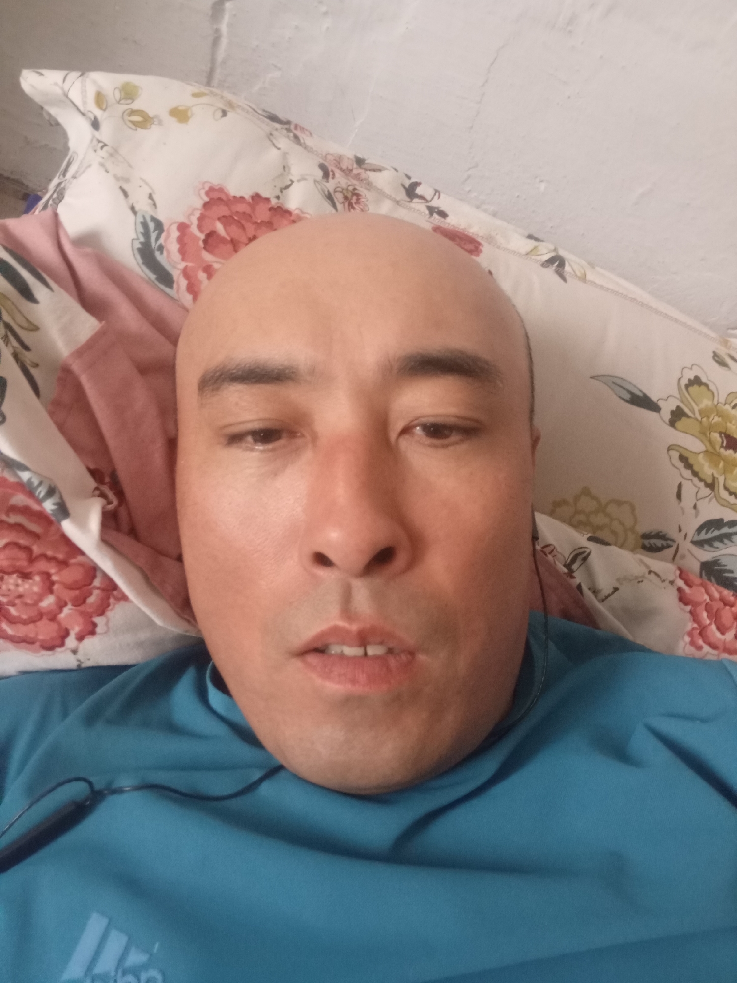 The image is a close-up, selfie-style photograph of a bald man lying down. He is wearing a blue t-shirt with an Adidas logo visible on the lower left. A black cord for earbuds is draped across his chest and neck. The man is lying on a pillow with a floral pattern featuring large pink and yellow flowers. A pink cloth is also visible behind his head. The background appears to be a textured, light-colored wall. There is no indication of the city of Baetov, Kyrgyzstan, in the image.