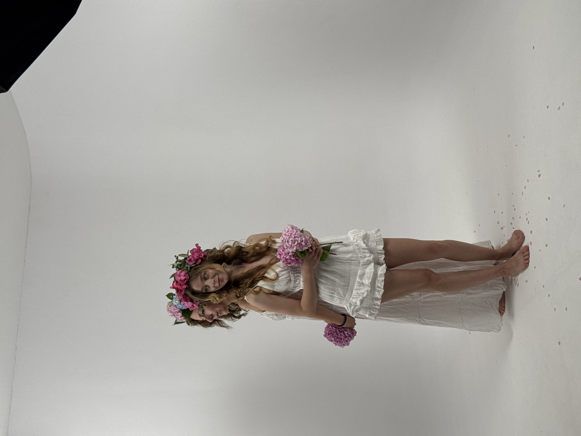 In a studio with a white backdrop, two women stand side-by-side, one partially obscuring the other. Both wear white dresses and flower crowns. The woman in front, facing the viewer's left, has curly brown hair and is holding a bouquet of pink flowers. The woman behind her, facing the viewer's right, has darker hair and is also holding a bouquet of pink flowers. Small pink petals are scattered on the white floor.