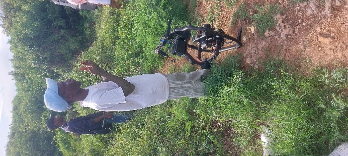 The scene depicts an outdoor, vegetated area within Dar es-Salaam, Tanzania, under an overcast sky. In the foreground-center, a young male of dark complexion, wearing a light blue baseball cap, a white t-shirt, and light-colored striped pants, stands in profile, facing right. His hands are raised in front of him, palms facing each other with fingers slightly bent. To his left and slightly behind him, another young male, dressed in a dark jacket and dark cap, stands partially obscured by vegetation, facing away from the viewer. On the far right, a third individual, mostly cropped by the frame, is visible wearing light-colored clothing. Resting on a reddish-brown dirt path in the foreground-right is a professional-grade camera mounted on a black stabilization gimbal system, equipped with a tripod base, indicating film or video production activity. The ground transitions from dense green grass and weeds on the left to the dirt path on the right. The background is dominated by a thick expanse of green bushes and small trees.