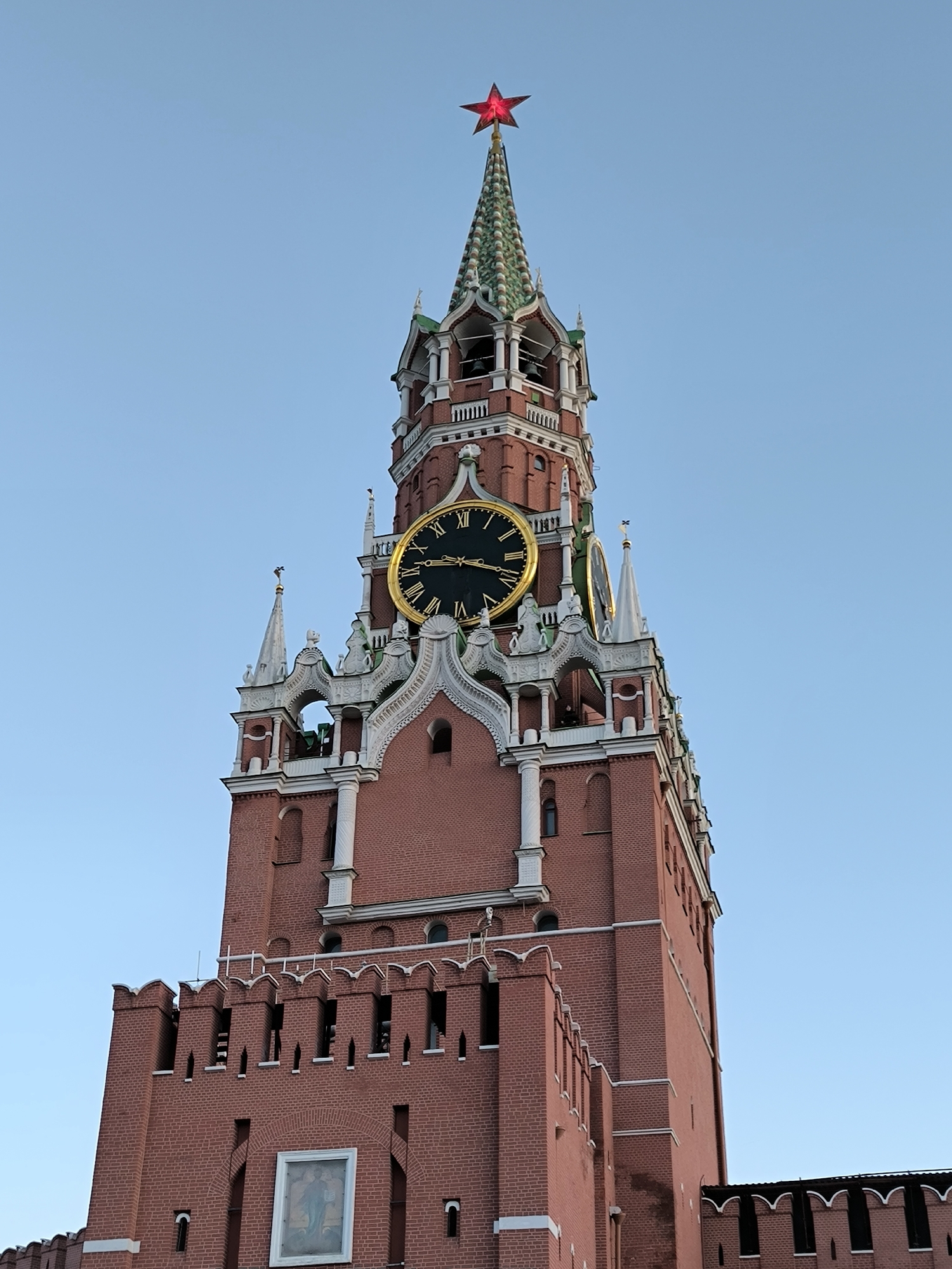 The image shows a close-up, low-angle view of the Spasskaya Tower in Moscow, Russia. The tower, made of red brick, features a large clock with Roman numerals and a golden frame. Above the clock, the tower narrows to a green, conical roof topped with a red, illuminated star. The sky behind the tower is clear and blue. The lower portion of the image reveals the crenellated battlements of the tower and a framed bas-relief image on the brickwork. The scene is devoid of people or any visible activity.