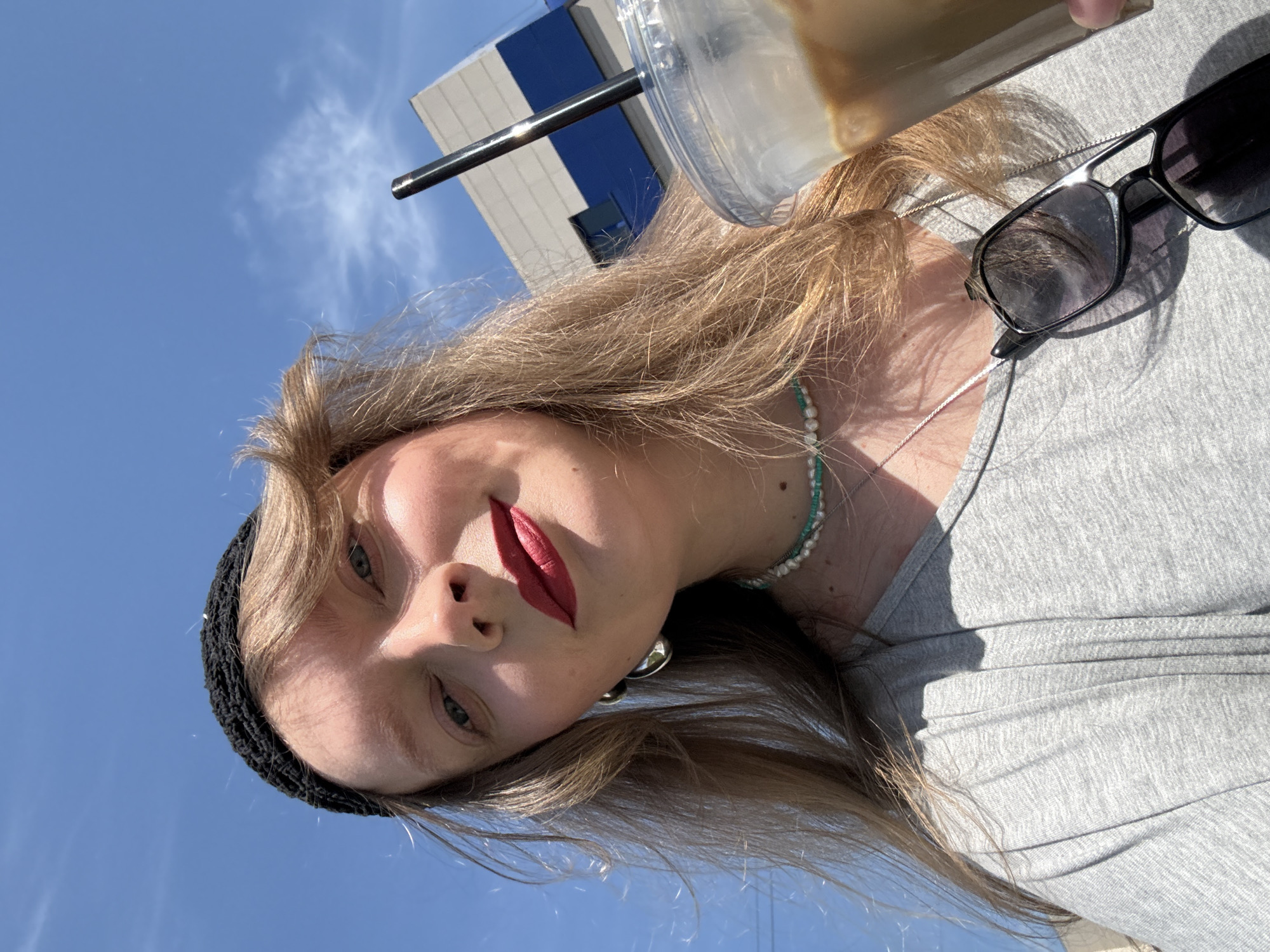 A woman is holding an iced coffee in front of her face. She is wearing a grey shirt and black knit cap. There is a building with blue and white panels in the background. The location is the city of Izhevsk, Russia. Sunglasses are next to her. She is wearing a necklace.