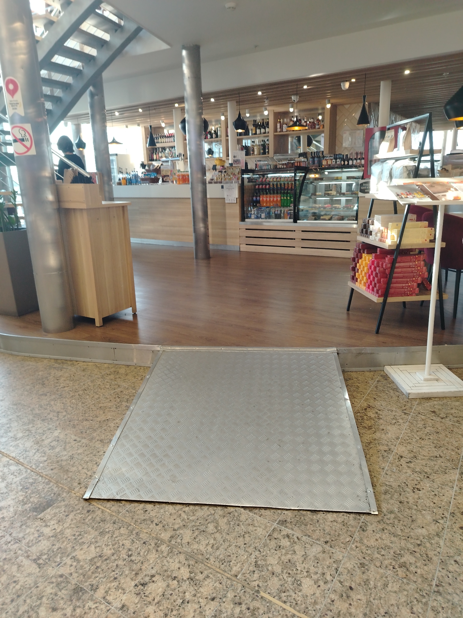 A wide-angle shot shows an indoor commercial space, possibly a cafe or dining area.
In the foreground is a diamond-plated metal ramp. It is positioned to bridge a slight elevation change. The floor surface is a speckled, light brown tiled pattern.
In the midground, the floor transitions to a wood-like material, indicating a different zone within the space. A supporting structural column is visible on the left, with a cylindrical metal base and a no-smoking sign. There's a wooden cabinet attached to the column's base.
A cafe or restaurant is set up in the background with a counter, displays, and food items, including tiered shelving with what appears to be packaged foods. The lighting is bright and airy, suggesting natural light sources. Bottles of alcohol are on a shelf behind the counter.
The location is Khimki, Russia.