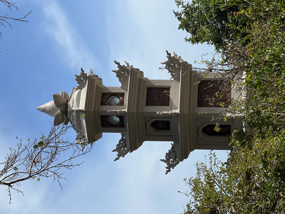 A tall, multi-tiered architectural structure, possibly a pagoda or tower, points toward a bright blue sky. The structure is off-white, with decorative elements including lotus-like finials and intricate carvings. Two circular windows or openings are visible. Branches of trees with sparse foliage frame the building on the left, while dense green trees are on the right. The image has been rotated, making the structure appear sideways.