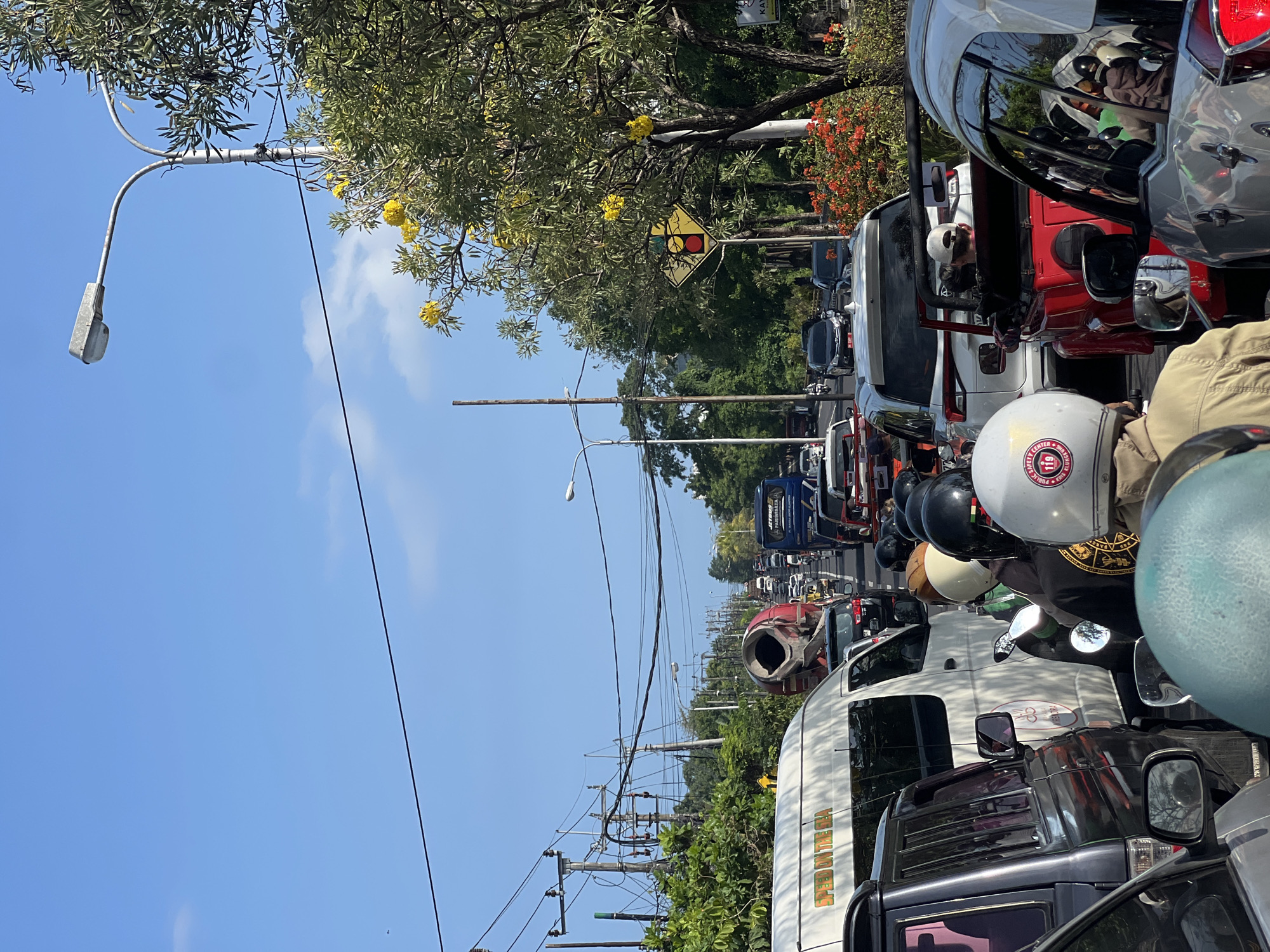 The image shows a busy street scene in Denpasar, Indonesia. A line of cars, vans, trucks, and motorcycles are stuck in heavy traffic. A red cement mixer is among the vehicles. People wearing helmets are on motorcycles. Power lines run along the side of the road. Trees with yellow flowers line the street. A street light and a traffic sign are visible. The sky is blue with scattered clouds.