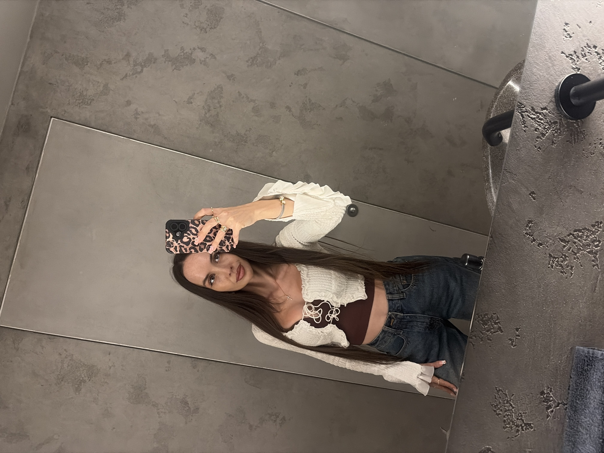A young woman with long, dark hair takes a mirror selfie in a bathroom. She is wearing a white, ruffled, long-sleeved crop top over a dark brown bralette, and blue jeans. She is also wearing several rings and a bracelet. The bathroom has grey, textured walls and a large mirror. A sink and faucet are visible in the lower part of the mirror's reflection. The scene is indoors, likely in Saint Petersburg, Russia, given the provided context.