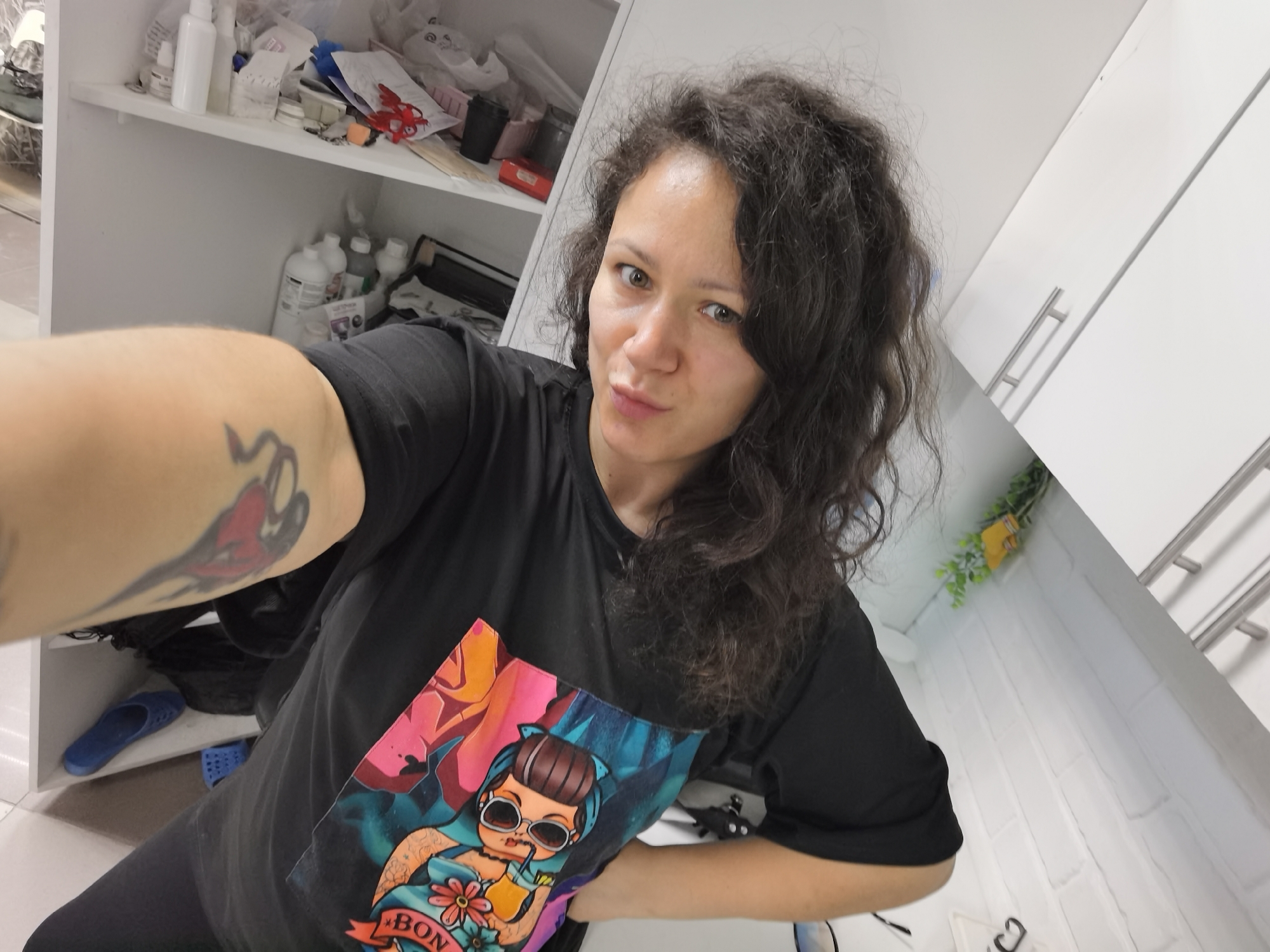 A woman with dark, curly hair and light skin is taking a selfie in a room with white walls and cabinets. She is wearing a black t-shirt with a colorful graphic design featuring a cartoon character with sunglasses and a drink. She has a tattoo on her left arm. In the background, there are shelves with various items, including bottles and papers. There is also a blue slipper on a shelf. The overall impression is that of a casual, indoor setting. The image does not contain any specific visual information that would allow for the identification of the location as Chaikovsky, Russia.