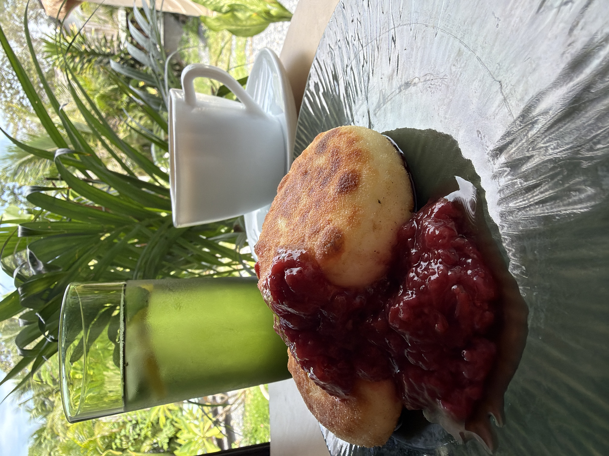 A close-up, eye-level shot shows a breakfast spread on a wooden table set against a backdrop of lush greenery, suggesting an outdoor or open-air dining setting, potentially in Ko Samui District, Thailand. In the foreground, a dark green textured plate holds two golden-brown syrniki (cheese pancakes) topped with a generous dollop of vibrant red berry jam. To the left of the plate, a tall glass filled with a green beverage, possibly iced tea or a cocktail, contains ice cubes and lemon slices. In the background, a white ceramic coffee cup and saucer sit to the right, with a white liquid, likely coffee, visible inside. The background is dominated by out-of-focus tropical foliage and palm leaves, hinting at a warm, humid climate and a natural setting, consistent with a location in Ko Samui, Thailand. The lighting suggests it is daytime, with natural light illuminating the scene. There are no people visible in the image.