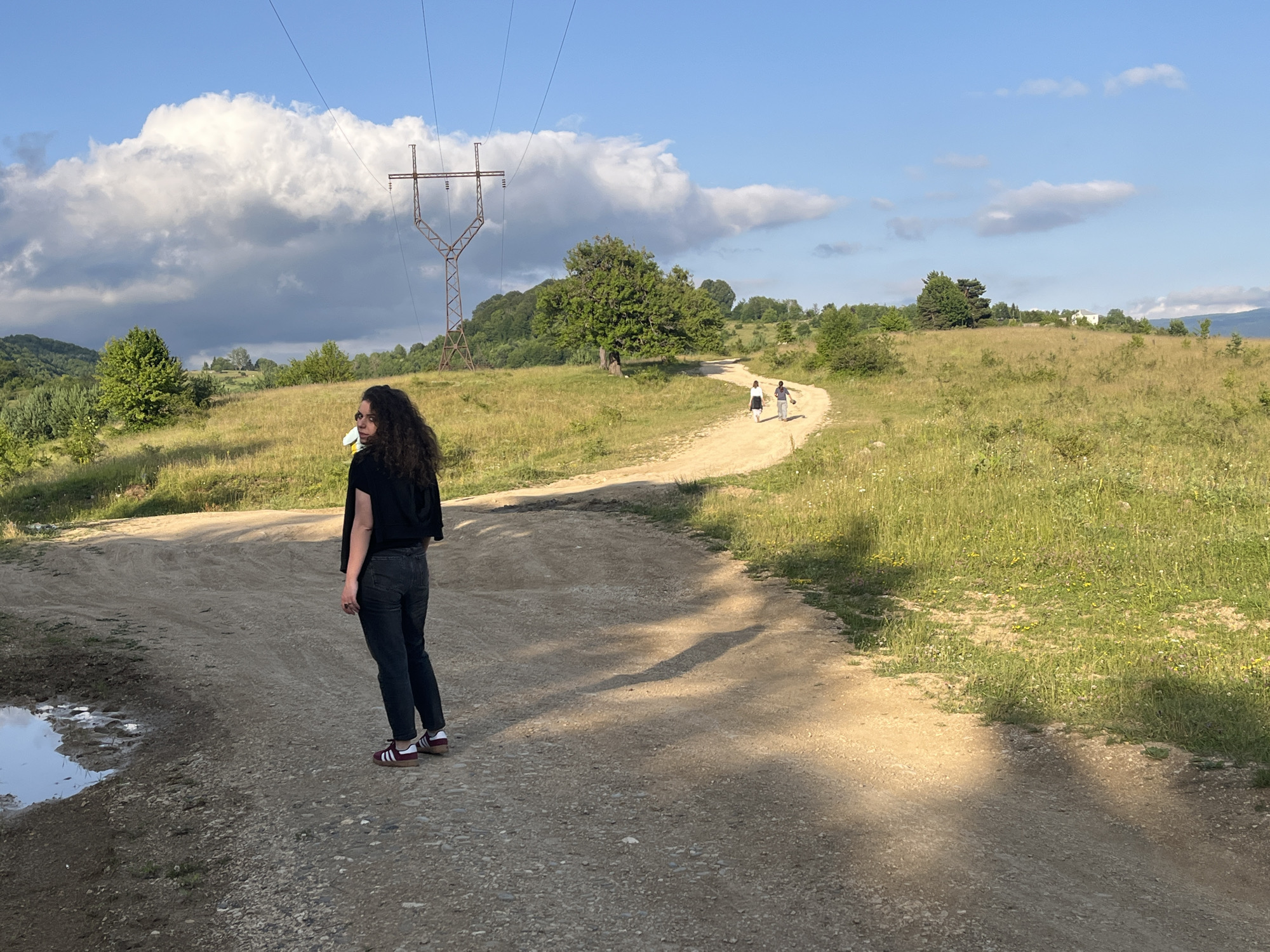 In Nikortsminda, Georgia, a young woman with dark, curly hair stands on a dirt road, facing away from the camera and looking over her left shoulder. She is wearing a black shirt and dark pants, with red and white sneakers. To her left, a muddy puddle reflects the sky. In the distance, two figures walk along the winding dirt road, which meanders through a grassy, hilly landscape. Towering electrical pylons and lines are visible against a bright blue sky filled with large, white clouds. Trees and foliage dot the hillsides, and a white building is faintly visible in the distance. The overall scene depicts a rural, outdoor environment on a sunny day.
