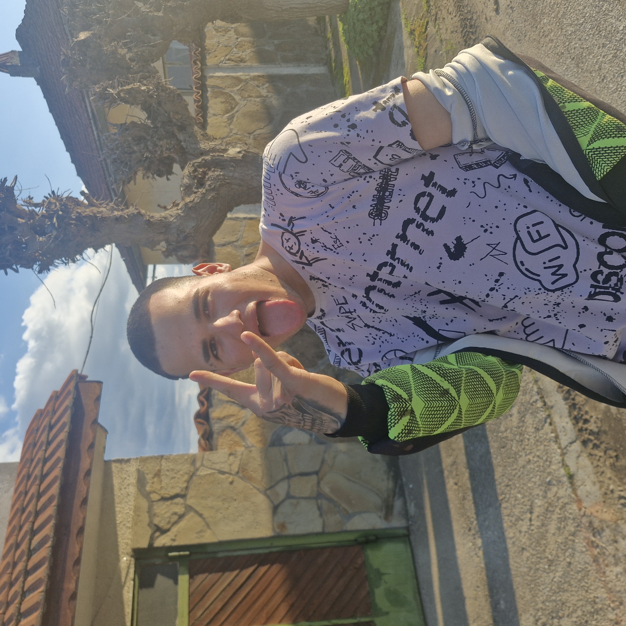 A light-skinned male with a shaved head, wearing a graphic-print white t-shirt with a white jacket over it and a lime green arm, is sticking his tongue out and making a "peace" sign with his left hand. He is standing on what appears to be a concrete sidewalk. In the background is an old building with a brick roof and a stone wall, and a tree with bare branches. The photograph was taken in Hirevo, Bulgaria.