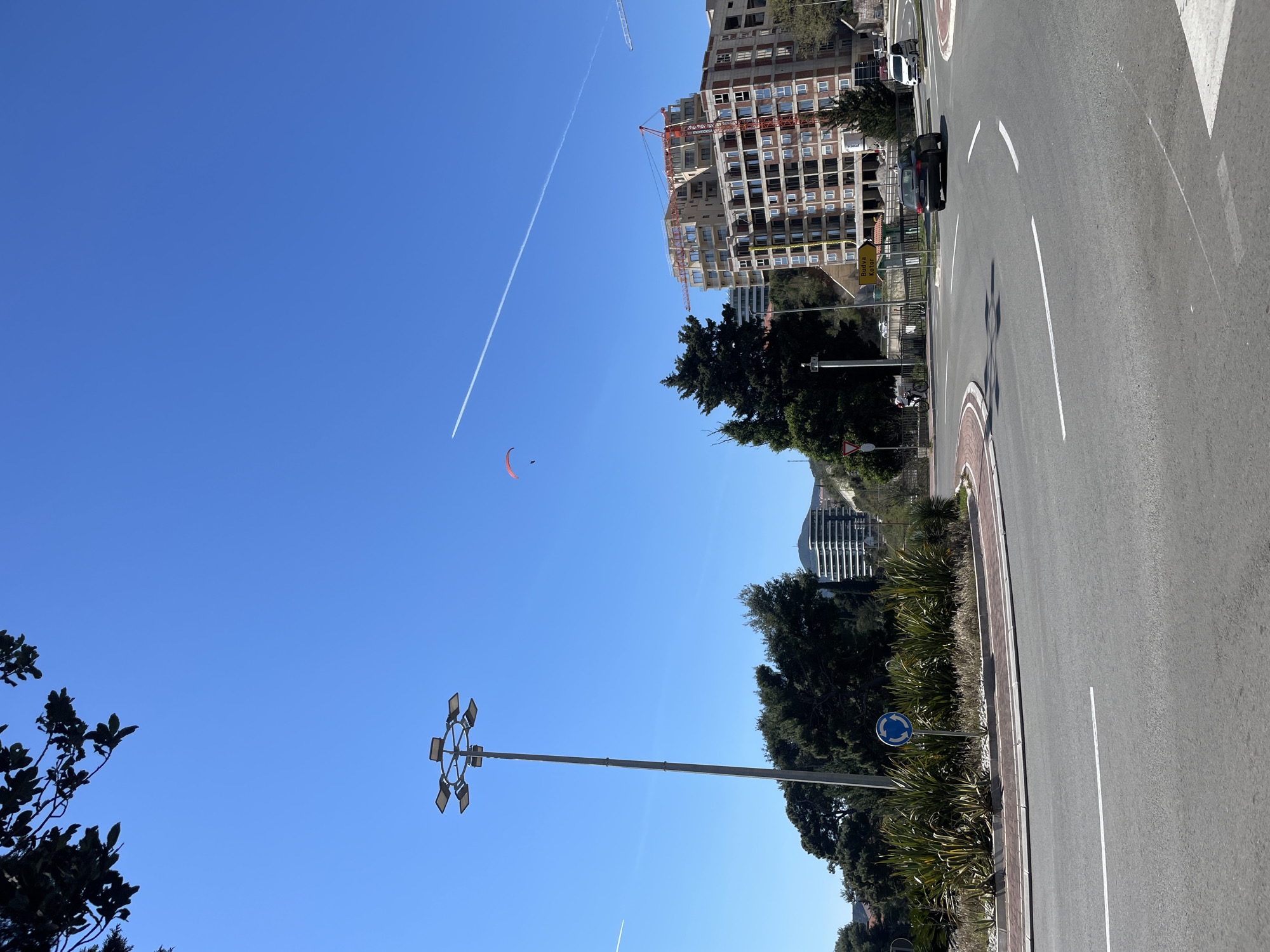 The image depicts a street scene in Rafailovići, Montenegro on a sunny day. The frame is dominated by a road with a roundabout and directional markings. Along the periphery of the road, there is vegetation including trees and shrubs. Behind the greenery, several buildings are visible, including a large structure under construction with a crane. In the sky, a paraglider is soaring, along with contrails from an aircraft. A tall street light is positioned along the roadside. Cars can be seen on the road.