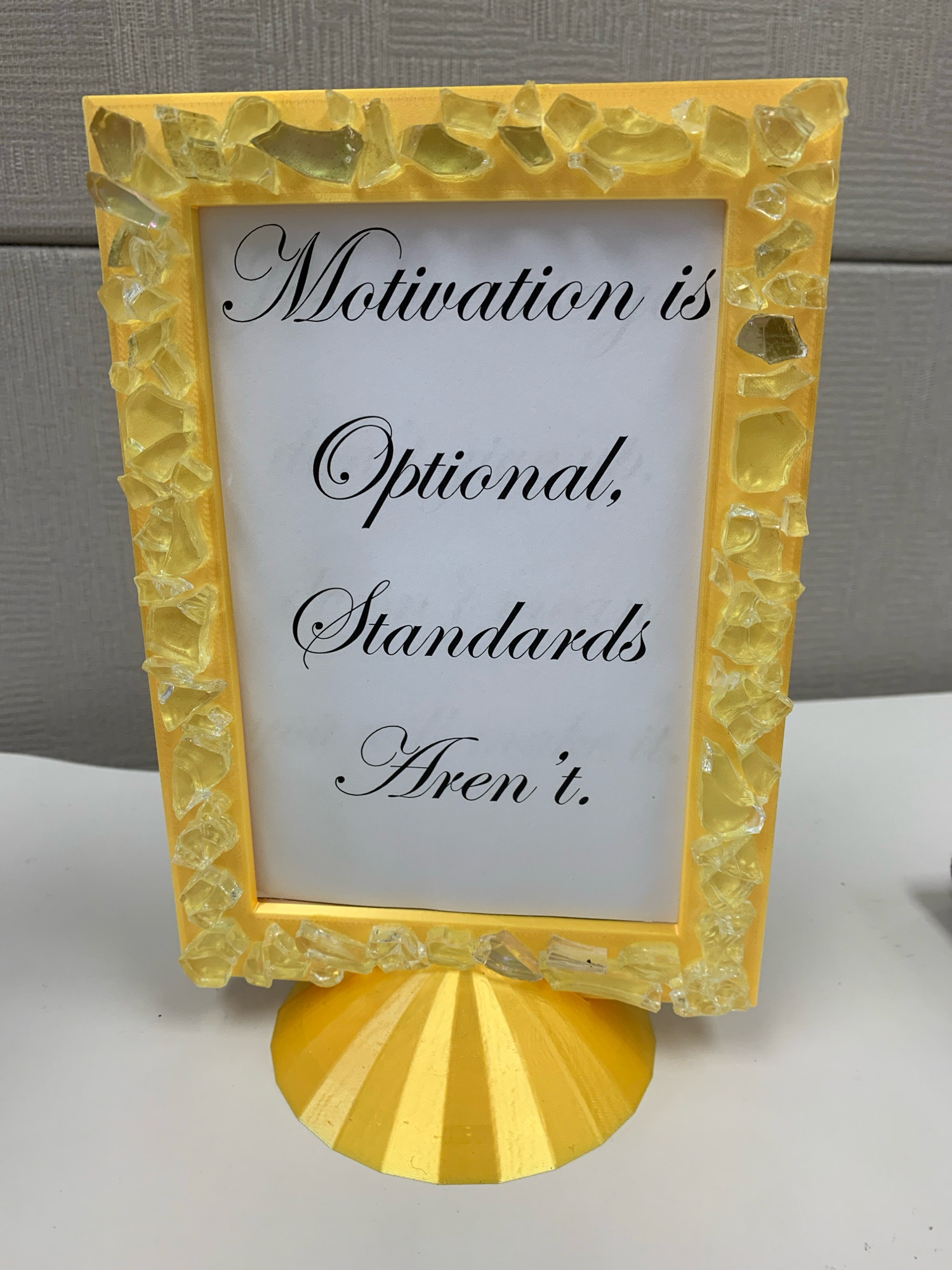 A framed motivational quote stands on a white surface. The frame is yellow and decorated with small, translucent yellow pieces. The quote, written in a black script font, reads: "Motivation is Optional, Standards Aren't." The frame is supported by a decorative yellow stand. The background appears to be a grey, textured wall. The image does not provide any information about its location in Brampton, Canada.