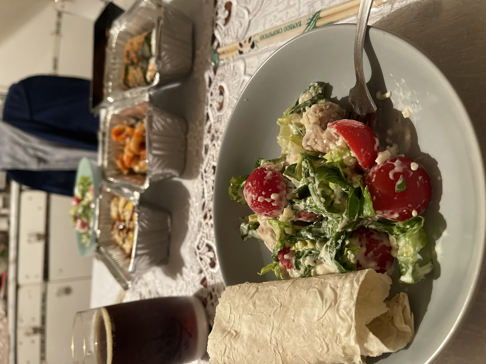 A close-up shot shows a meal laid out on a table, likely in a domestic setting in Khimki, Russia. The foreground is dominated by a grey plate holding a salad with chopped lettuce, cherry tomatoes, and what appears to be pieces of chicken or fish, all coated in a creamy dressing. A rolled flatbread rests on the left side of the plate. To the right of the plate, a fork is positioned, and a pair of chopsticks, branded "BAMBOO CHOPSTICKS", are visible. In the background, several foil containers hold takeaway food, and a glass of dark liquid, possibly beer, is on the left. A person, partially visible in a blue jacket over a grey shirt, is seated in the background. The table is covered with a white, intricately patterned tablecloth. The overall scene suggests a casual meal, potentially a takeaway dinner.