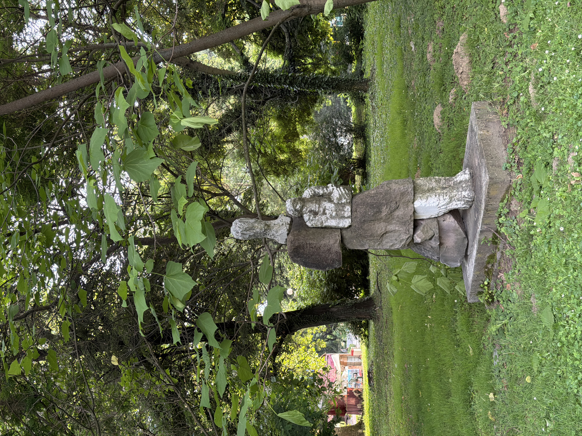 The image depicts a stone sculpture situated in a grassy park area. The sculpture, composed of several stacked stones of varying shapes and sizes, resembles a stylized human figure. It stands on a concrete base. The surrounding environment is lush and green, with trees and foliage providing a natural backdrop. Sunlight filters through the leaves, creating dappled lighting. In the background, a building with a red roof and some outdoor seating can be seen. The ground is covered in grass and small plants. The overall impression is of a tranquil outdoor setting, likely a public park or garden.