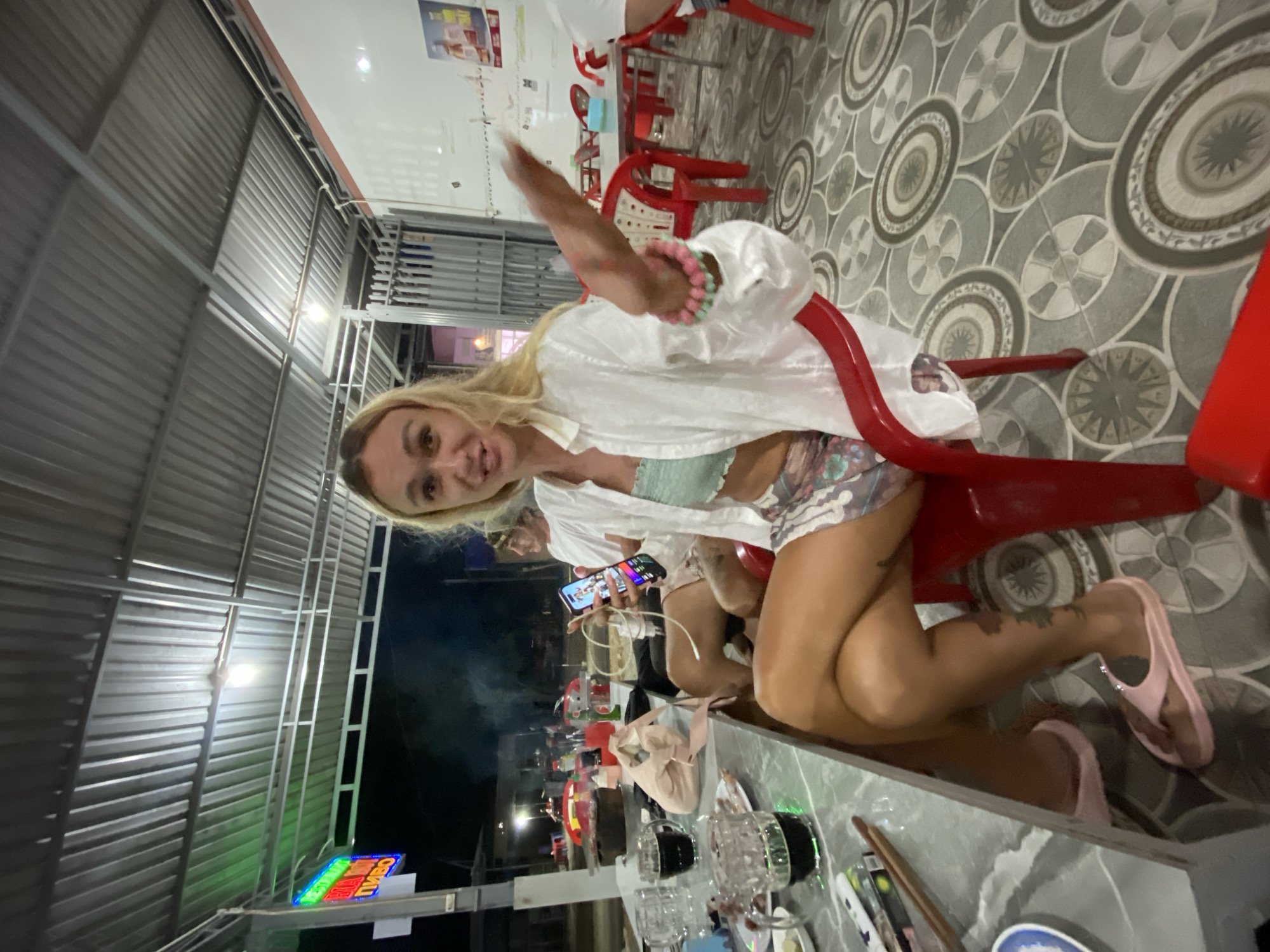 The image depicts a nighttime scene in a semi-outdoor dining establishment located in Phu Thuy Ward, Vietnam. The central figure is a blonde-haired female, appearing to be a young adult, seated on a red plastic chair. She is wearing a translucent white long-sleeved shirt over a light green tube top, floral shorts, and pink flip-flops. Her left shin displays a circular tattoo. She wears multiple beaded bracelets in pink and light green on her right wrist. She holds an illuminated smartphone in her left hand, while her right hand is raised with an open palm, gesturing towards the viewer. Her mouth is open, indicating speech or a reaction.
At the gray marble-patterned table in front of her, two large glass mugs containing a dark liquid and a smaller glass with a green liquid are visible. A white plate holds food remnants, and chopsticks, a pack of Marlboro cigarettes, and a lighter are also on the table. A crumpled beige cloth is present. Further down the table, a black cooking pot, likely for hotpot, emits smoke.
Behind the central figure, a dark-haired male is seated, engaged with a smartphone. Other individuals are partially visible at adjacent tables. The dining area is covered by a corrugated metal roof supported by metal beams, with an overhead light fixture providing illumination. To the left, a neon sign displays red, green, and yellow text, including the Cyrillic word "ПИВО" (PIVO - beer). The floor is adorned with a repeating pattern of concentric circles and stars in gray, white, and brown tiles. A white wall with posters is visible on the right side. Red plastic chairs and metal-framed tables are standard throughout the establishment. Beyond the covered area, a dark street is visible with distant artificial lighting.