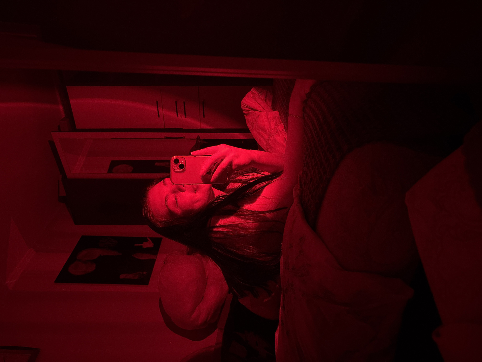 A person is taking a selfie in a room illuminated by red light. The person is holding a smartphone with multiple camera lenses visible on the back. A bed with pillows and blankets is on the right side of the frame. A cabinet or dresser is visible in the background. There is a framed picture on the wall. The location is indicated as Perm, Russia.