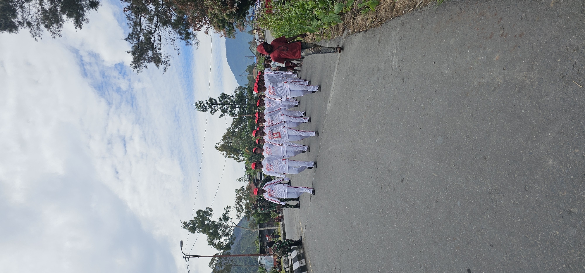 In Puncak Jaya, Indonesia, a group of approximately 15 individuals, predominantly women, are participating in a parade on an asphalt road. They are uniformly dressed in white outfits with red accents and red caps. Several individuals have "WORK OUT" printed in red on their shirts, and one person in the front row has the number "13" on their shirt. They are marching in formation, facing the camera.
To the left of the marching group, two individuals in military camouflage uniforms are standing near a black and white striped barrier. Behind them, several other people are visible sitting or standing. In the background, there are trees, houses, and mountainous terrain.
To the right of the parade, a young male wearing a red shirt and patterned pants is walking away from the camera. The road is lined with vegetation on the right side. The sky above is partly cloudy with patches of blue. The overall scene suggests an organized event or procession taking place in an outdoor, possibly rural or semi-urban, setting.