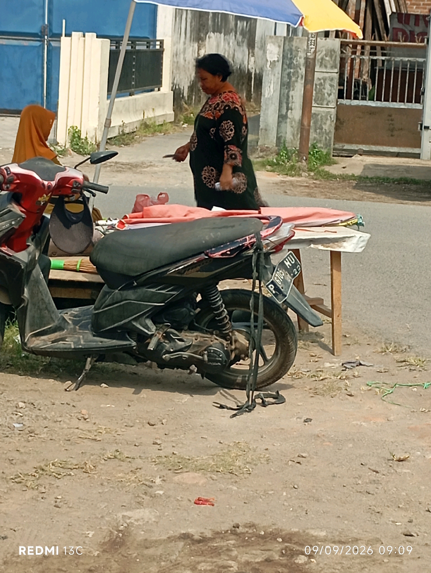 A woman in a floral dress stands next to a motorbike, with a woman in an orange hijab seated on the back of the motorbike. The scene appears to be on the side of a road in Wuluhan, Indonesia. A small table with a pink and white cloth is set up in front of the woman in the floral dress, suggesting a roadside stall. The background shows a blue gate and a fence. The ground is dirt and gravel. The image is dated 09/09/2026 at 09:09.