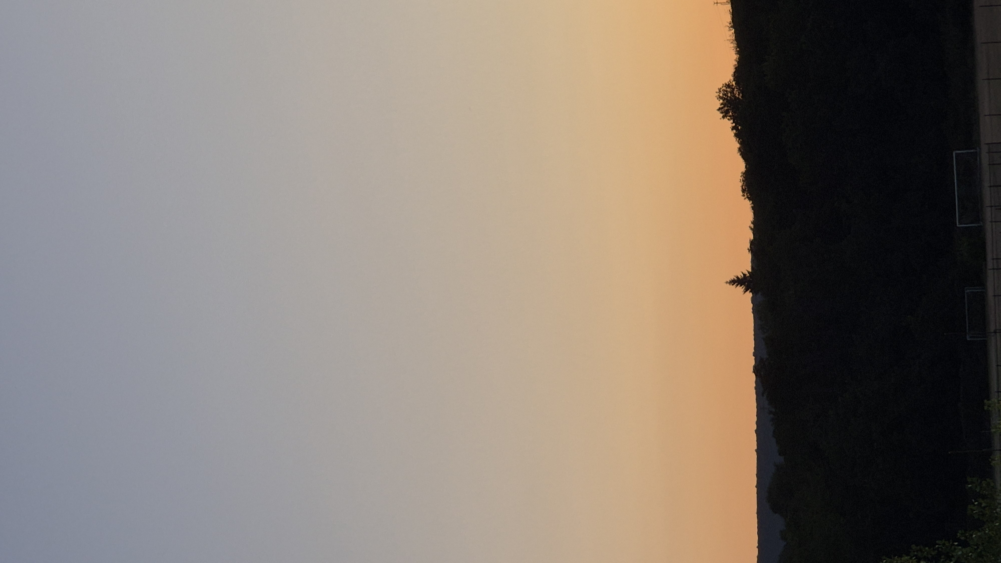 This is a vertical, medium-shot, outdoor photograph of a sunset over a forested area. The sky transitions from a pale blue at the top to a warm orange near the horizon. A silhouetted line of dark trees forms the mid-ground, with a single, tall evergreen tree standing out against the sky. In the foreground, two silhouetted soccer goals are visible on a field. The image appears to be taken from a slightly elevated perspective, looking towards the west. No people or animals are visible in the frame. The location is identified as Lovech, Bulgaria.
