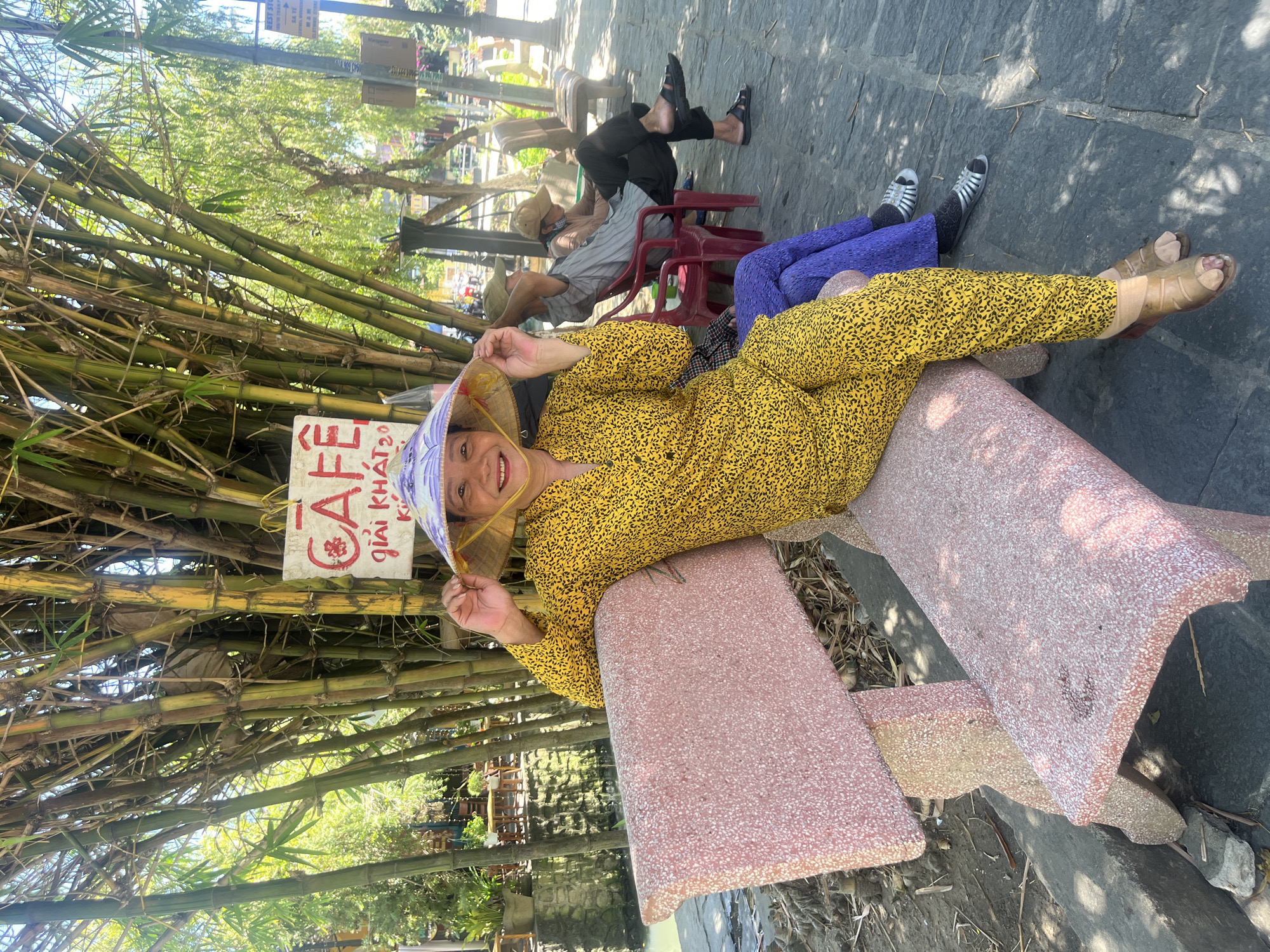 The image captures a vibrant, sunny daytime scene in what appears to be an outdoor cafe or resting area, likely in Hội An Ward, Vietnam. The photo is taken with a vertical orientation, showing several individuals relaxing.
In the foreground, a woman lies comfortably on a pink and white speckled stone bench, smiling broadly towards the camera. She is dressed in a bright yellow long-sleeved shirt and matching pants, both featuring a small, dark patterned design. She wears a traditional Vietnamese conical hat (nón lá) adorned with purple and white patterns, holding it lightly with her left hand. Next to her, the lower legs of another person are visible, clad in bright purple pants and silver sandals worn over black socks.
Behind the main subject, a dense wall of tall green bamboo stalks forms a natural boundary on the left side of the frame. Attached to the bamboo is a small white sign with prominent red text. Further in the background, several other people are visible, also relaxing. One person sits on a red plastic chair, wearing a light-colored striped shirt and a hat, while another, positioned further back, wears dark pants and sandals. The ground is a dark grey stone path, marked by dappled sunlight and shadows, indicating a clear, sunny day. Green trees and foliage are visible beyond the bamboo.
The white sign prominently displays "CAFE" in large red letters, with smaller red text below it that reads "GIẢI KHÁT," which translates to "Refreshments" in Vietnamese. The overall mood of the image is one of leisure and happiness, set in a pleasant, tropical outdoor environment.