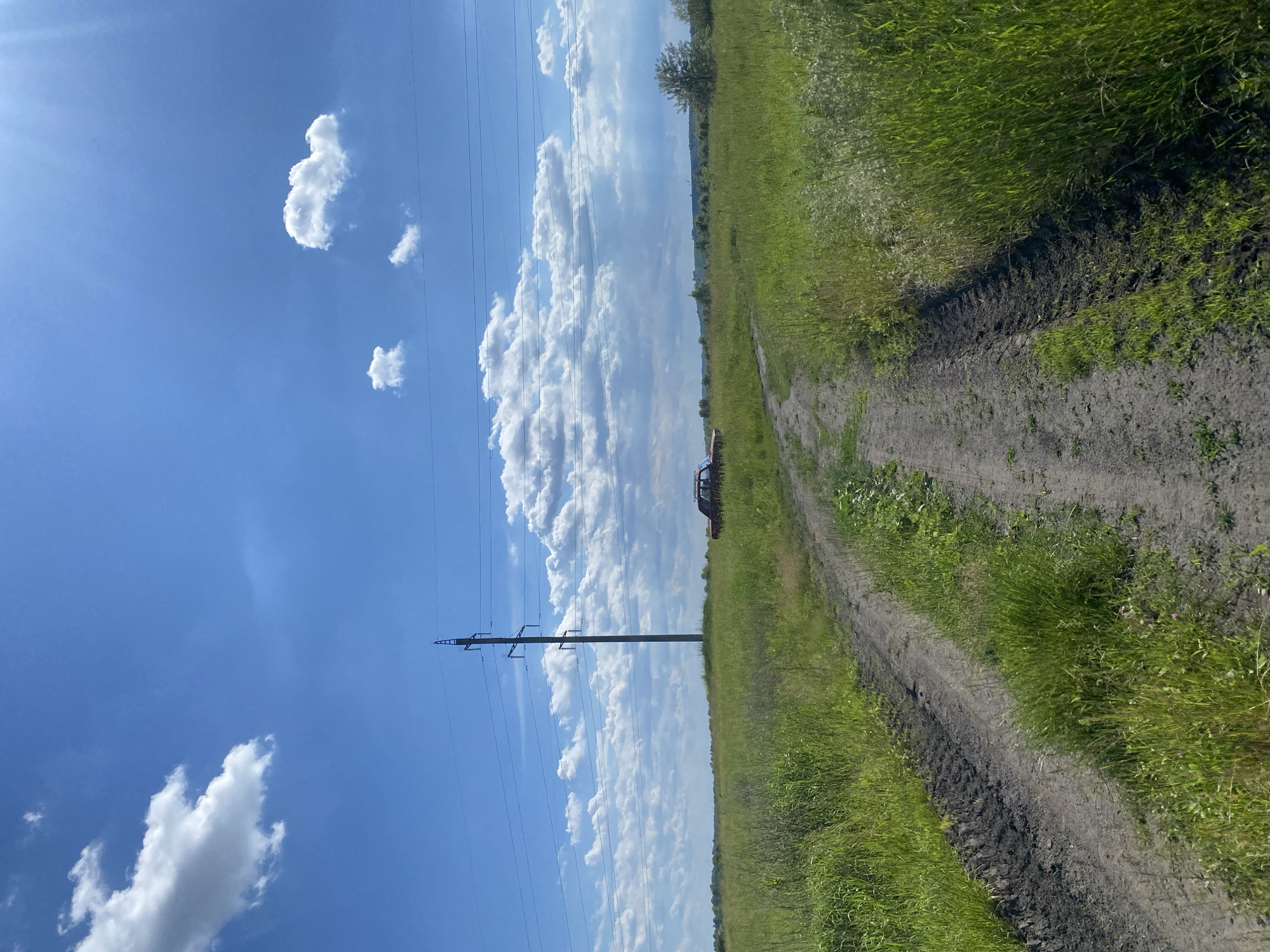 The image shows a dirt road cutting through a grassy field under a bright blue sky with scattered white clouds. A single, tall power line pylon stands in the middle distance, with several power lines stretching across the sky. To the right of the pylon, a rusty red car is parked in the field. The road appears to have tire tracks, suggesting recent use. In the far distance, a treeline is visible, indicating the edge of the field. The overall scene suggests a rural or semi-rural environment.