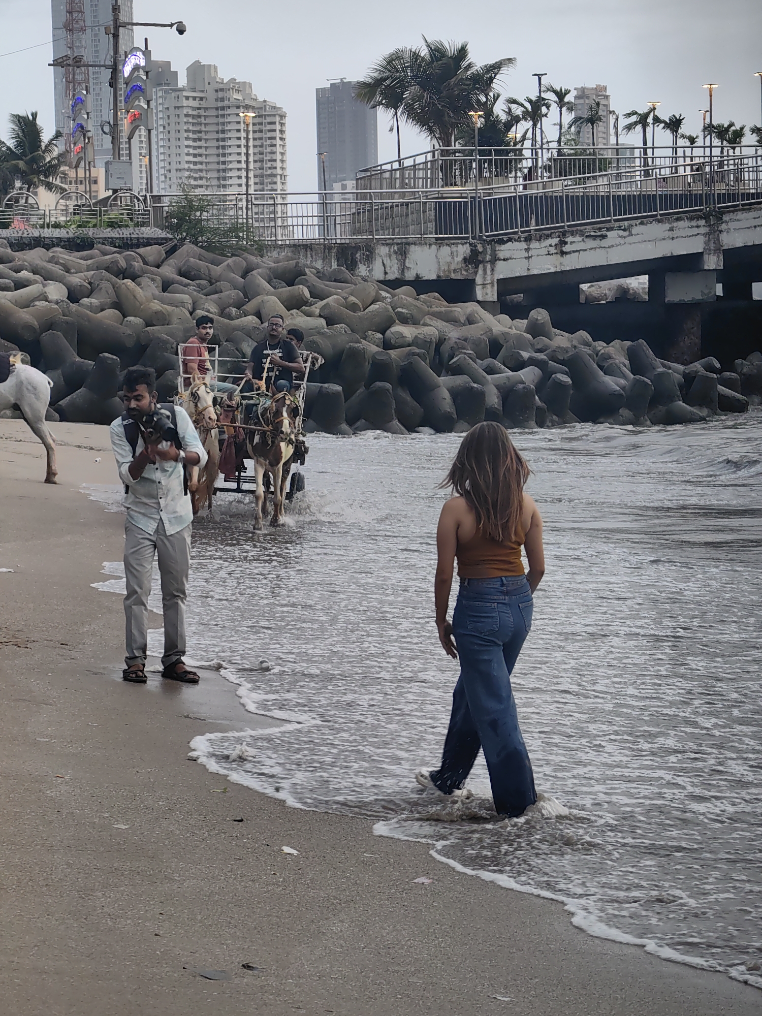 This image depicts a scene on a beach in Mumbai, India, likely near Marine Drive, given the iconic tetrapods and the cityscape in the background. The sky is overcast, suggesting either early morning or a cloudy day.
In the foreground, a man wearing a light blue collared shirt and grey pants stands on the wet sand, holding a small black dog. He is facing left and appears to be interacting with the dog, which he holds cradled in his arms. He is wearing sandals.
Further back, a horse-drawn cart is visible on the shallow water's edge, moving from left to right. Two men are seated in the cart. The driver is in the front, and another man is seated behind him. Another person is partially visible behind the second man.
To the right of the cart, a woman with long brown hair is walking into the water. She is wearing a brown tank top and blue flared jeans. Her feet are submerged in the waves, and she is facing away from the camera, looking towards the ocean.
The background features a shoreline lined with large concrete tetrapods, commonly used as coastal defense structures. Beyond the tetrapods, a bridge or elevated walkway is visible, with a railing and lampposts. In the far background, the Mumbai skyline is dominated by several high-rise buildings, including a prominent skyscraper with a crane on top. Palm trees are also scattered throughout the scene. The water is a murky grey-brown, typical of urban coastlines.