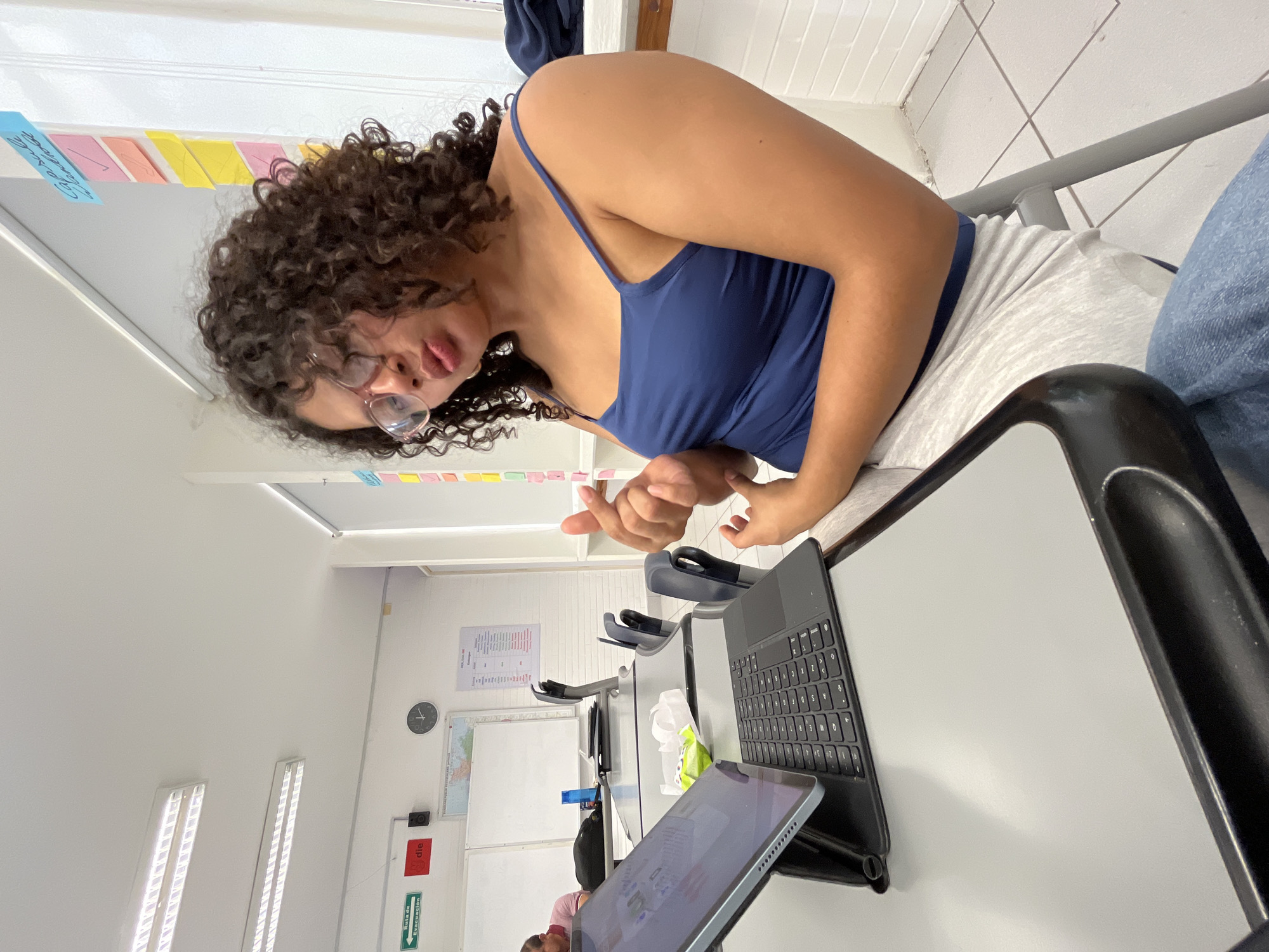 A young woman with curly dark hair, wearing glasses and a blue tank top, is sitting at a desk in a classroom. She is looking down at a tablet and keyboard. The classroom has white walls with fluorescent lighting and colorful sticky notes affixed to a whiteboard. A world map and other charts are visible on the wall. Other desks and chairs are visible in the background. This image was taken in the city of Puebla, Mexico.
