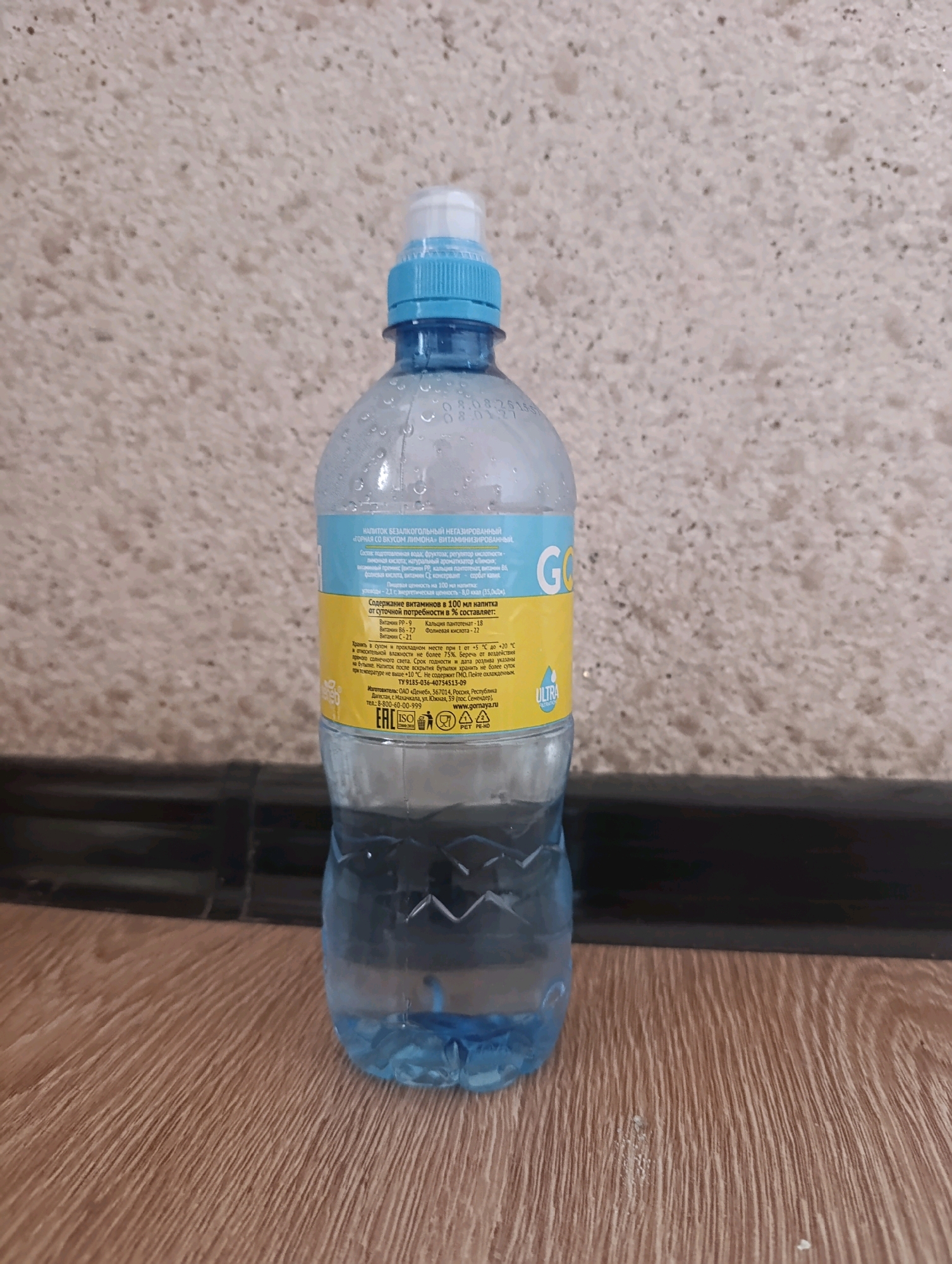 A clear plastic bottle filled with water and condensation is standing upright on a wooden surface. The bottle has a blue cap and a label with blue and yellow text. The background is a textured wall.