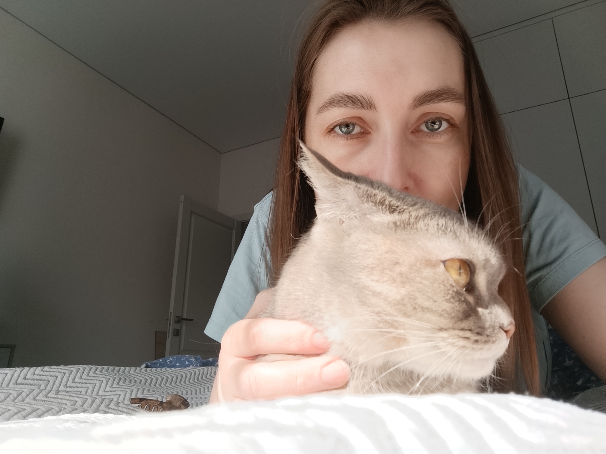 A woman is in a room with a light-gray cat. The woman is looking at the camera with a slightly downward angle. She is wearing a light-green shirt. Her hair is long and brown. The cat is light-gray with yellow eyes and is partially obscured by the woman's hand. The cat is held close to her face. The bed is covered with a gray and white zigzag-patterned cover. The room appears to be a bedroom, with light-gray walls and a door visible in the background. A pair of brown eyeglasses are on the bed in front of the bed cover. It is not possible to determine from the image whether it was taken in the city of Chaikovsky, Russia.