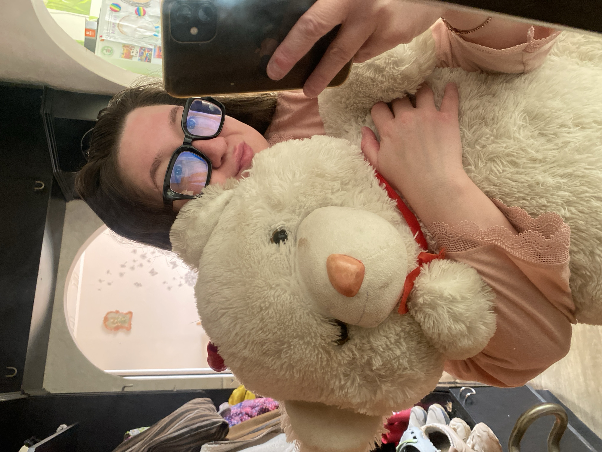 An indoor photograph, likely a mirror selfie, depicts a female individual holding a large cream-colored teddy bear within a domestic setting in Chaikovsky, Russia. The individual, visible from the upper chest upward, wears black-framed glasses and a light pink, long-sleeved top with lace detailing on the cuff of the left sleeve (viewer's right). Her dark hair is partially visible. Her lips are slightly pursed. She holds a dark-colored smartphone, equipped with a visible dual-lens camera module, in her right hand (viewer's left), positioned to capture the image in the reflective surface. Her left hand (viewer's right) is wrapped around the torso of the large teddy bear. The bear features dark, small eyes, an orange-tan nose, and a red ribbon tied around its neck.
The background, visible as a reflection, shows an interior space. To the left, a dark furniture unit, possibly a wardrobe or shelving, contains folded striped fabric, other garments, and light-colored footwear. A golden, curved handle is affixed to a dark surface in the lower left foreground. Centered behind the individual, an arched doorway leads to an adjacent room with light-colored walls. On this wall, a light orange decorative relief or plaque is mounted, accompanied by several small, dark, irregular decals to its right. Above the archway, a dark furniture piece displays a silver U-shaped hook. To the right, a cluttered area, possibly a kitchen or storage space, is partially reflected, showing colorful items and various containers.