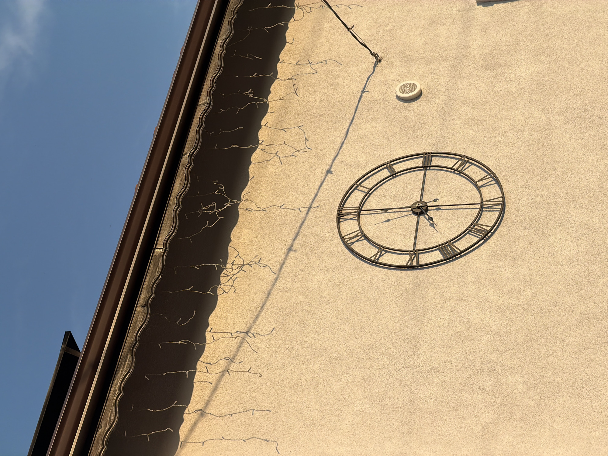 The image shows a close-up of the exterior wall of a building with a large, ornate clock featuring Roman numerals. The clock is mounted on a light-colored, textured wall. Above the clock, a brown gutter system runs along the edge of the roof. Icicle-like string lights hang from the gutter, casting shadows on the wall. The sky above is clear and blue. The scene is illuminated by warm sunlight, suggesting it is either early morning or late afternoon. The image was captured in Pazardzhik, Bulgaria, though no specific landmark is identifiable to pinpoint the exact location within the city. There are no people visible in the image.