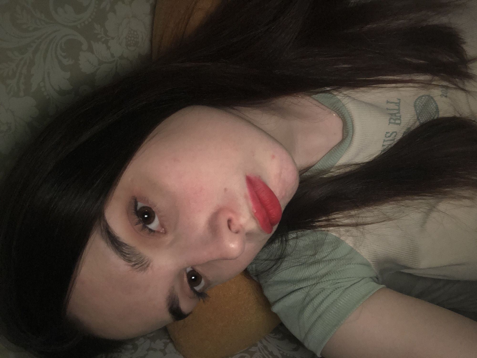 A woman with long, dark hair is lying down, possibly on a bed or sofa, with her head tilted towards the camera. She has fair skin, dark eyebrows, and is wearing red lipstick. Her t-shirt is light-colored with green sleeves and the words "TENNIS BALL" visible. The background appears to be a patterned wallpaper or fabric. There is a yellow pillow behind her head. The photo doesn't provide any information to place it in Noviy, Russia.