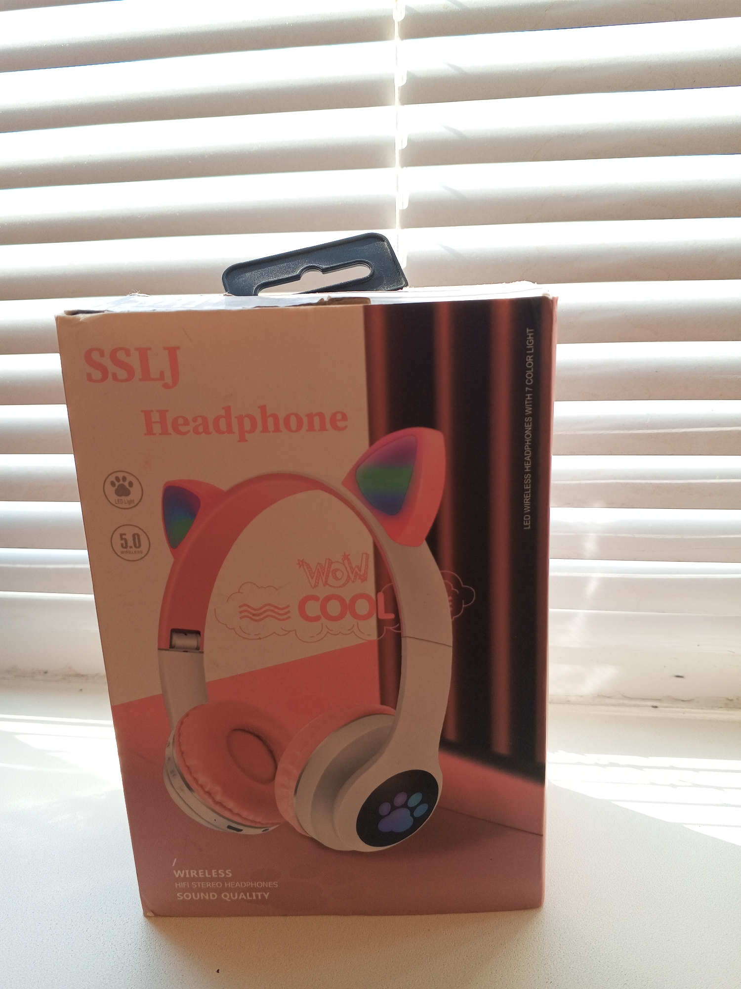 The image shows a pink and white box for SSLJ wireless headphones with cat ears. The box has a picture of the headphones, which are also pink and white, with colorful earcups and a paw print design on the side. Text on the box reads "SSLJ Headphone," "LED Light," "5.0 WIRELESS," and "WOW COOL." The box also advertises "WIRELESS HIFI STEREO HEADPHONES SOUND QUALITY" and "LED WIRELESS HEADPHONES WITH 7 COLOR LIGHT." The box is placed on a white surface in front of a window with white horizontal blinds. The scene is illuminated by natural light coming through the blinds, casting striped shadows on the surface. There are no people or any identifiable locations within the city of Chistopol, Russia, visible in the image.