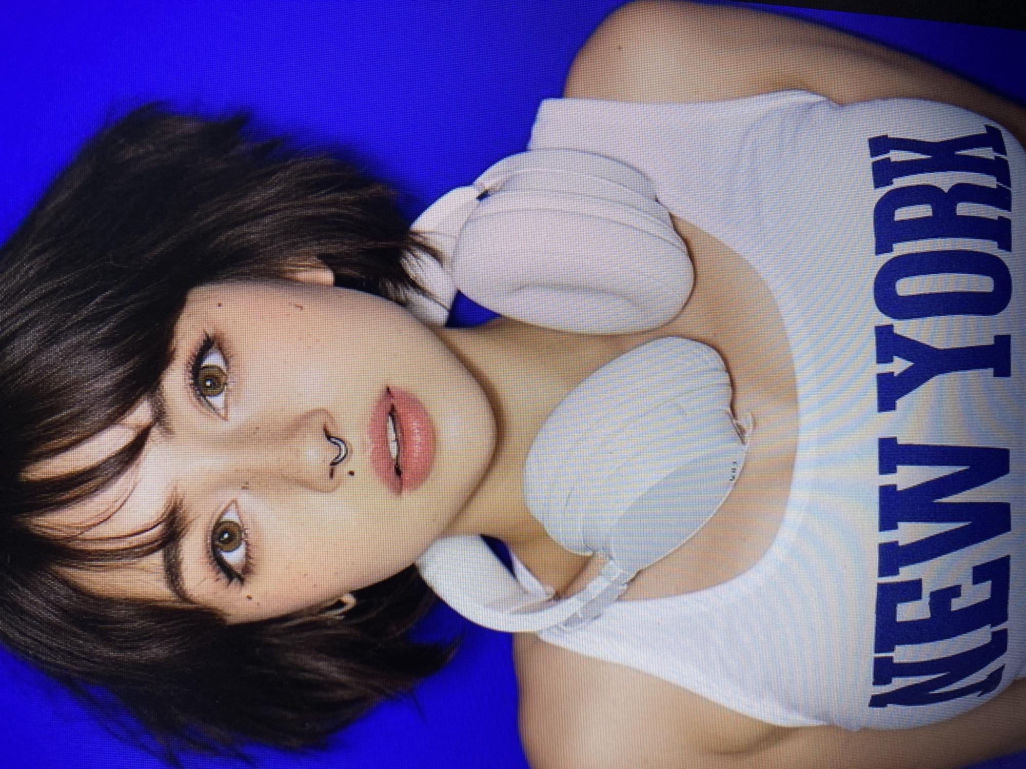 A young woman with dark, short hair and light brown eyes is pictured from the chest up. She has freckles on her face and a septum piercing. She is wearing a white tank top with "NEW YORK" printed in blue letters across the front, and white headphones around her neck. The background is a solid blue. The image appears to be a photograph. No location in Yerevan, Armenia, is discernible from the image.