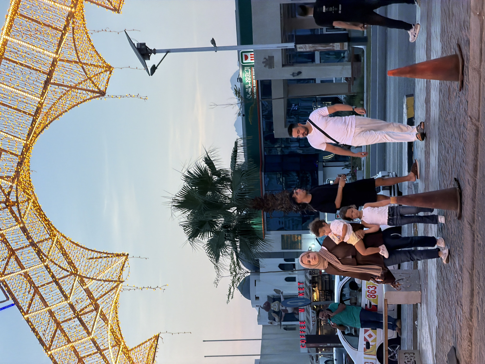 A family walks along a paved sidewalk in Sharm El Sheikh, Egypt, under a large, illuminated archway decorated with golden lights. A woman wearing a hijab and a brown abaya carries a toddler on her hip, while a young girl in a white tank top and jeans walks beside her. Behind them, a man in a white t-shirt and white pants walks with a black crossbody bag. In the background, palm trees and buildings are visible, including a structure with Arabic and English text that reads "NATIONAL BANK OF EGYPT." A traffic cone stands on the sidewalk in the foreground. The sky is clear and pale blue, suggesting it is late afternoon or early evening.