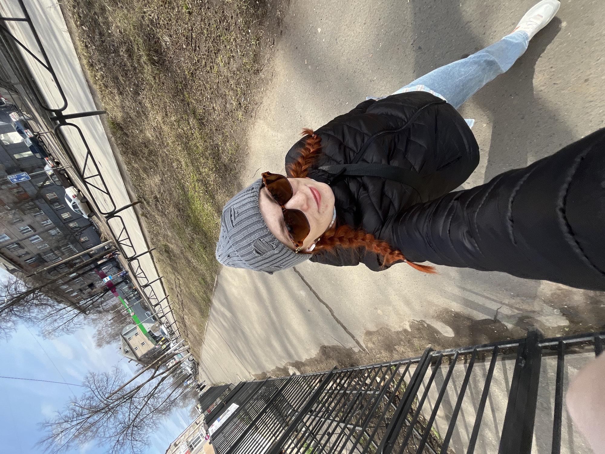 The image shows a selfie taken outdoors on a sunny day. The subject is a woman with red braids, wearing a grey beanie, brown sunglasses, a black puffer jacket, light blue jeans, and white sneakers. She stands on a concrete sidewalk next to a black metal fence.
Across the fence, there is a view of apartment buildings in the city of Chaikovsky, Russia, identified by typical Soviet-era architecture. Trees are visible, partially obscuring the buildings, and the sky is blue with some clouds. A grassy area is seen between the sidewalk and the buildings. The sidewalk appears to be slightly wet in places.