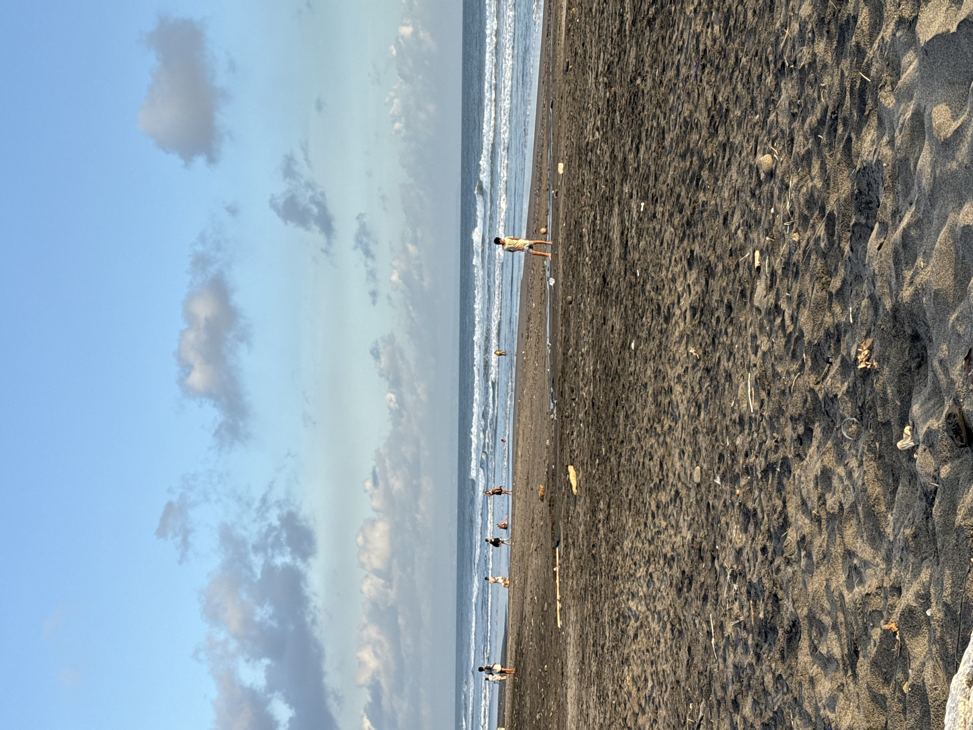 The image depicts a beach scene in an unknown city. The beach has dark sand. A few people are visible along the shoreline. The sky is blue with scattered clouds. The ocean has gentle waves. There's some debris scattered on the beach.