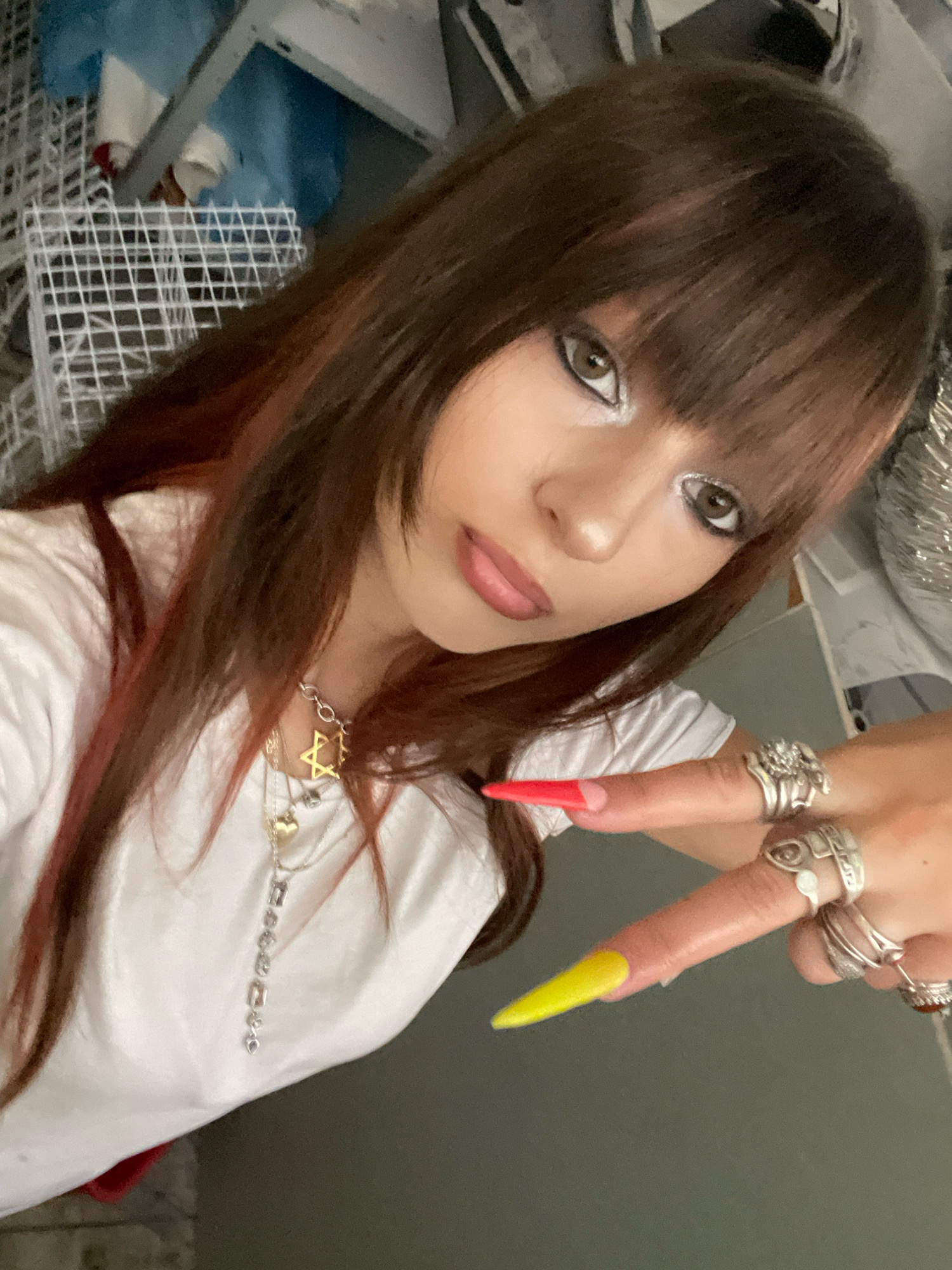 A young woman with brown hair with auburn highlights and bangs is taking a selfie. She is wearing a white t-shirt and multiple silver rings on her fingers, which have long, pointed acrylic nails in red and yellow. She is also wearing a silver necklace with star and heart pendants. Her makeup includes dramatic eyeliner and silver eyeshadow. The background appears to be an indoor setting with various objects, including what looks like storage shelves and possibly some laundry baskets. The location is not identifiable as Raanana, Israel, or any other specific city.