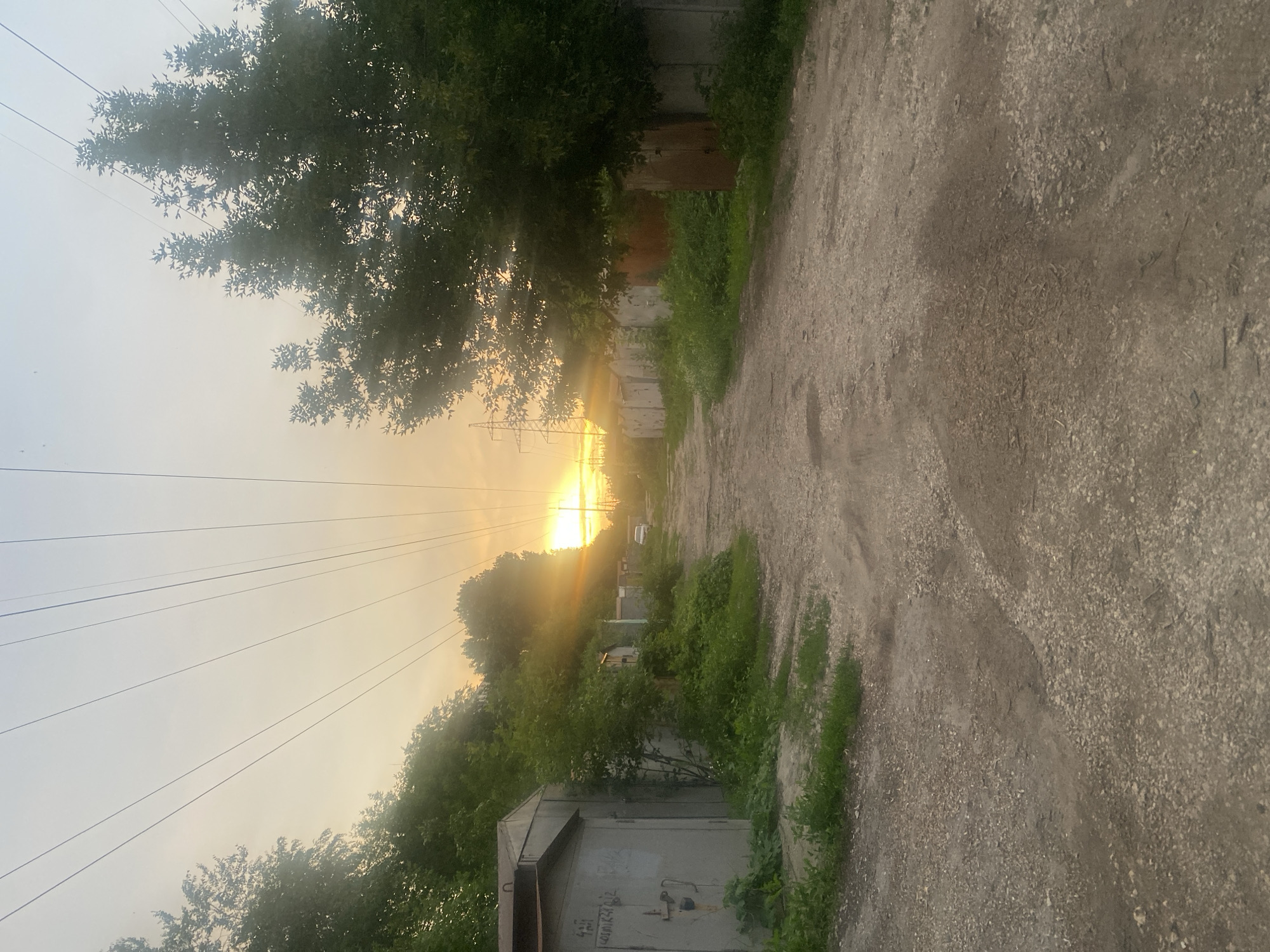 The image depicts a dirt road lined with concrete garages on both sides, leading towards a setting sun. The sun is low on the horizon, casting a warm glow and creating lens flare effects. Power lines stretch across the sky, converging towards the horizon. Lush green trees and vegetation are visible on either side of the road, partially obscuring some of the garages. A white car is parked in the distance. The overall impression is a slightly overgrown, somewhat neglected area, possibly a garage cooperative, common in many Russian cities, including Samara. The scene appears to be captured during the early evening, as indicated by the low angle of the sun and the soft, warm light.
