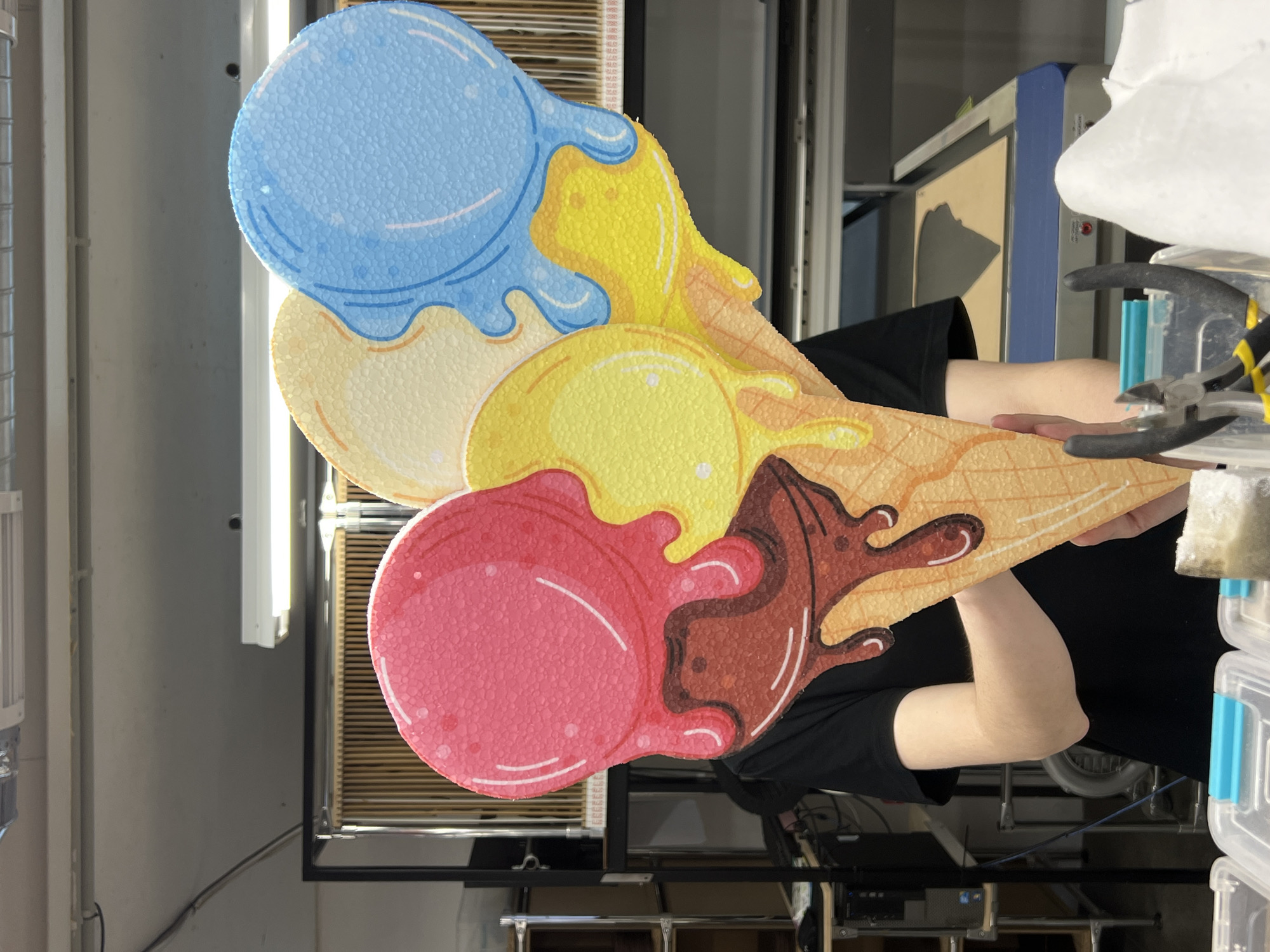 The image displays an indoor scene, likely a workshop or production area, captured sideways. A person, whose arms and torso are visible, holds a large, brightly colored cut-out decoration shaped like three scoops of melting ice cream on a waffle cone. The person wears a black short-sleeved shirt.
The ice cream decoration features a light blue top scoop, a yellow middle scoop, and a pinkish-red bottom scoop, all depicted with a melting effect and outlined with darker shades or white lines. The scoops and melting drips have a textured, foamy appearance, resembling expanded polystyrene or a similar lightweight material. The base of the decoration is a light brown waffle cone pattern, with brown drips flowing from the bottom scoop.
In the background, various industrial elements are visible. These include vertical metal supports or shelving units, horizontal fluorescent light fixtures, and what appears to be a blue and gray machine. To the right, a pair of pliers with yellow handles rests near clear plastic storage containers with blue latches. Further back, a white, irregularly shaped object and what looks like wooden slats within a frame are partially visible. The lighting is artificial, suggesting an indoor setting. There is no clearly legible text in the image.