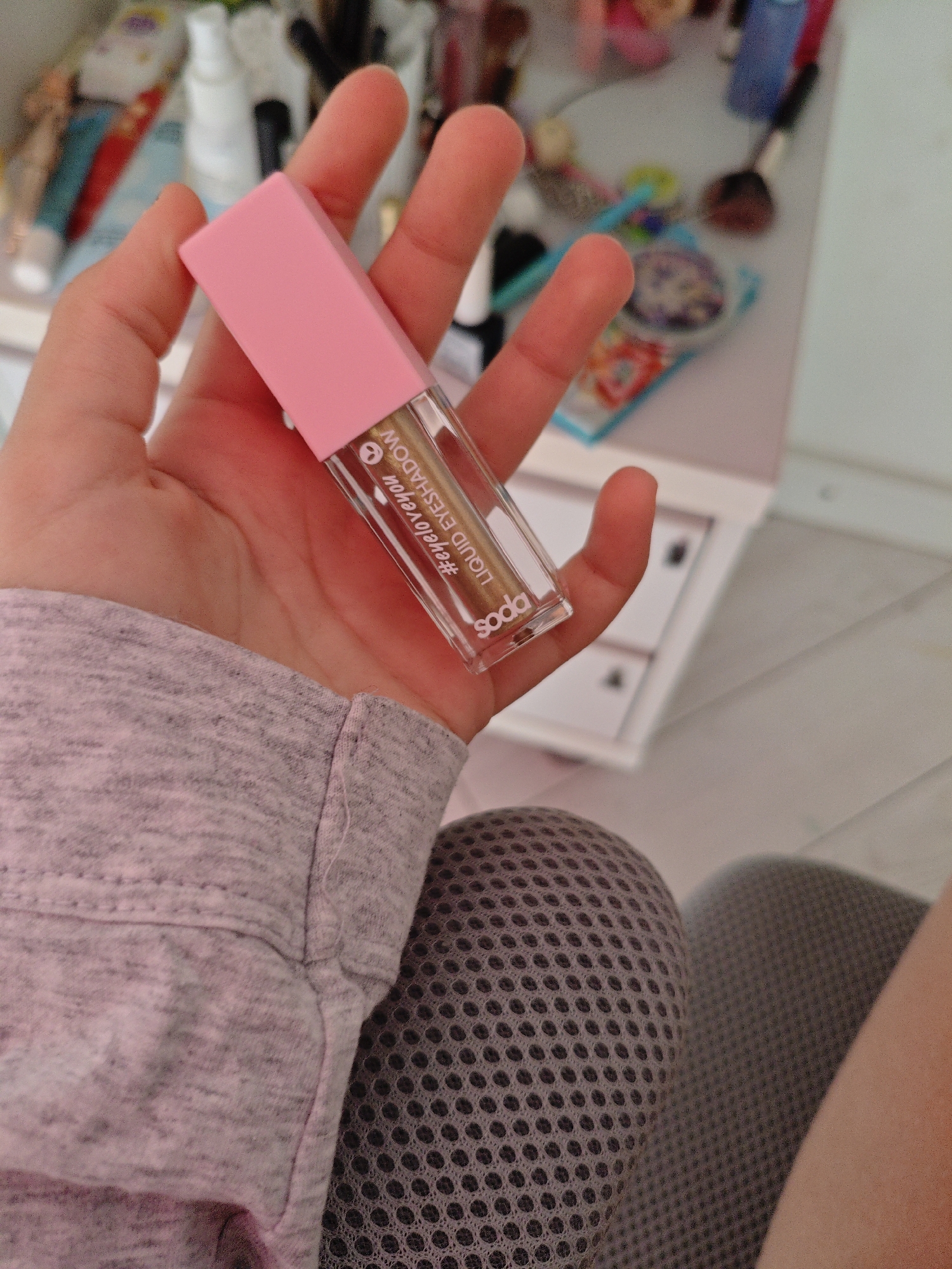 A person holds a pink and clear liquid eyeshadow tube with gold shimmer. The tube has text that reads "#eyeloveyou liquid eyeshadow" and "soda". The person's hand is in focus, and they are wearing a grey long-sleeved shirt. In the background, a blurred vanity with makeup products is visible. The scene appears to be an indoor setting, likely a bedroom or dressing area. There is no information that allows for the localization of this image in Трудовское сельское поселение, Ukraine.
