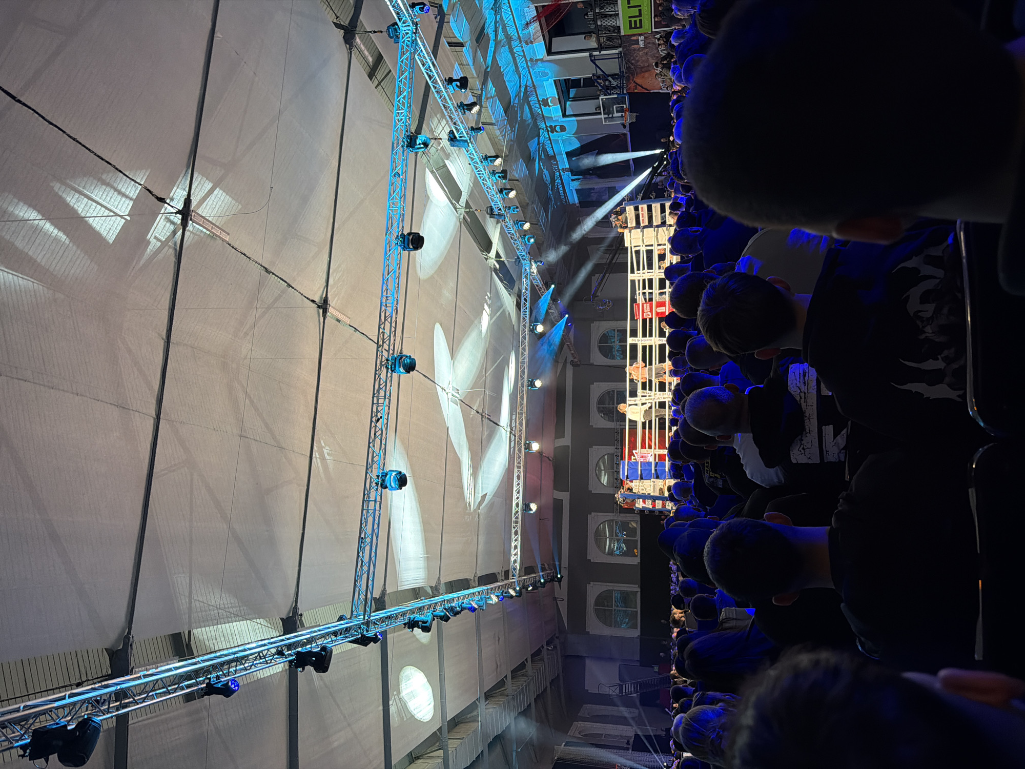 The image shows an indoor event, possibly a boxing match or a concert, based on the elevated platform in the center of the space surrounded by spectators. The venue has a high ceiling with a white fabric covering, possibly for acoustic or aesthetic purposes. A blue-lit metal structure, likely for lighting and sound equipment, spans the length of the room. Spectators are densely packed around the platform, facing the action. The city is Sofia, Bulgaria, based on the ELITE logo that says "Sofia, Bulgaria" on the right-hand side of the image.